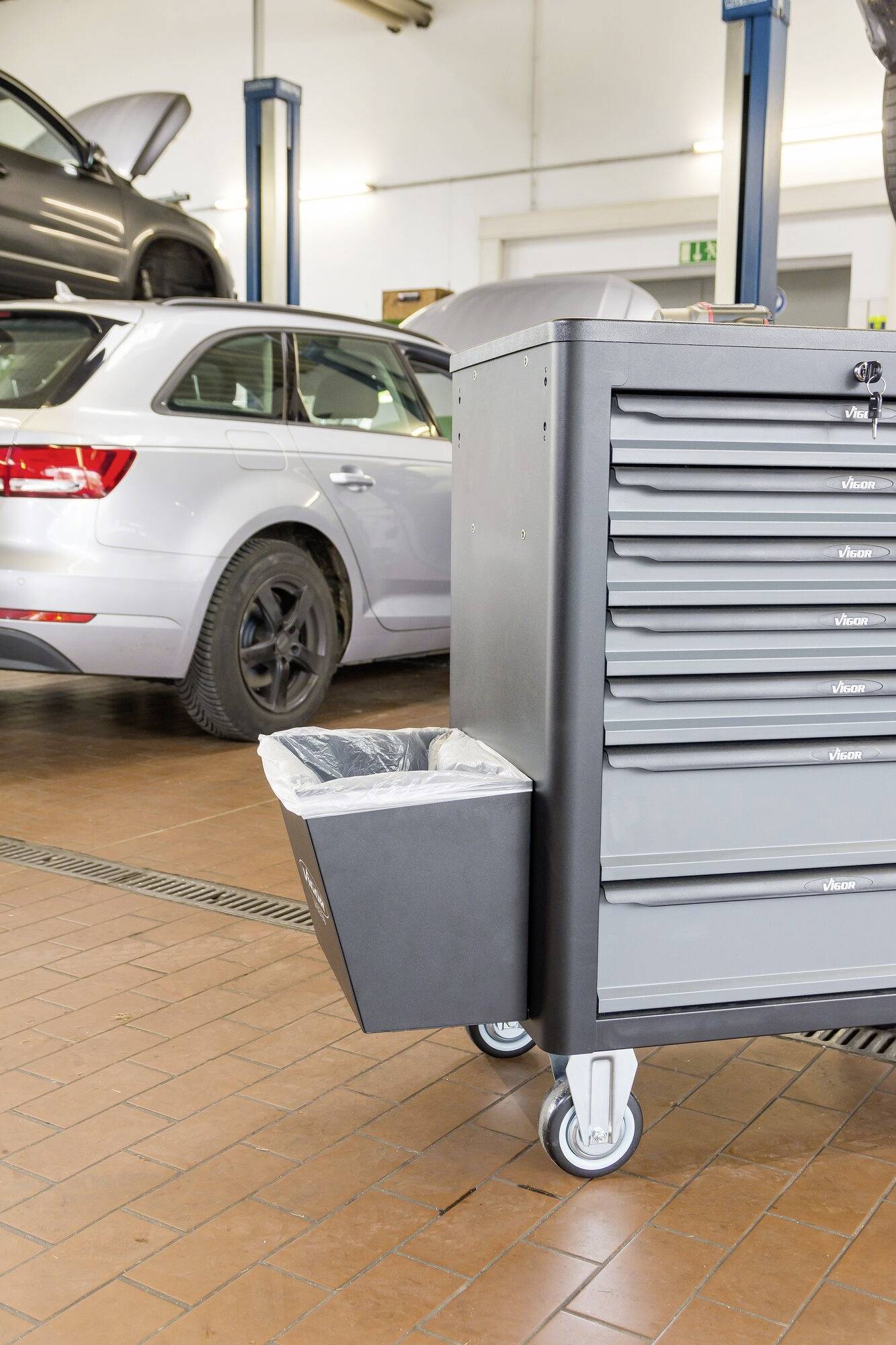 W warsztacie samochodowym na podnośniku stoi srebrny samochód. Na pierwszym planie znajduje się szary wózek narzędziowy z sześcioma szufladami.