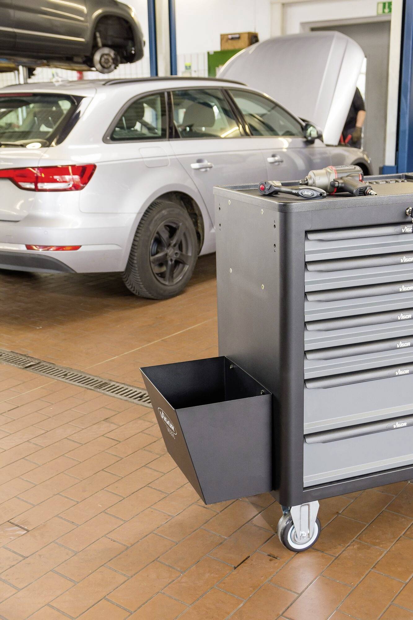 Szary samochód z otwartą klapą bagażnika stoi w warsztacie. Na pierwszym planie widoczny jest wózek narzędziowy z szufladami.