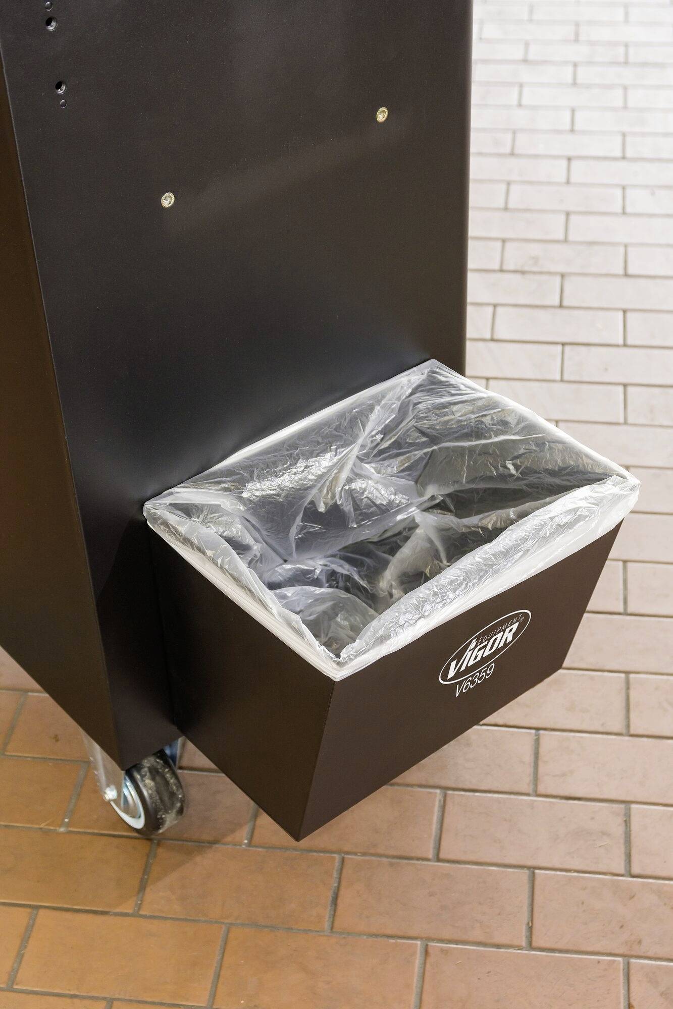 Czarny kosz na śmieci z przezroczystym workiem foliowym, stojący na kółkach. Na koszu widnieje napis 'VIGOR PACKS'. Podłoga wykonana jest z cegieł.