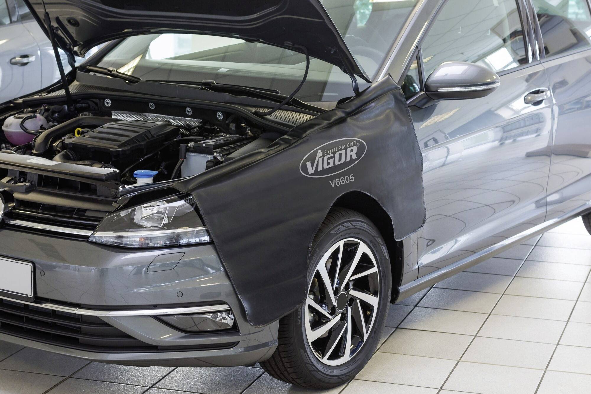 Szary samochód stoi w warsztacie z otwartą maską. Ochronny pokrowiec z napisem 'Vigor V6805' przykrywa błotnik.