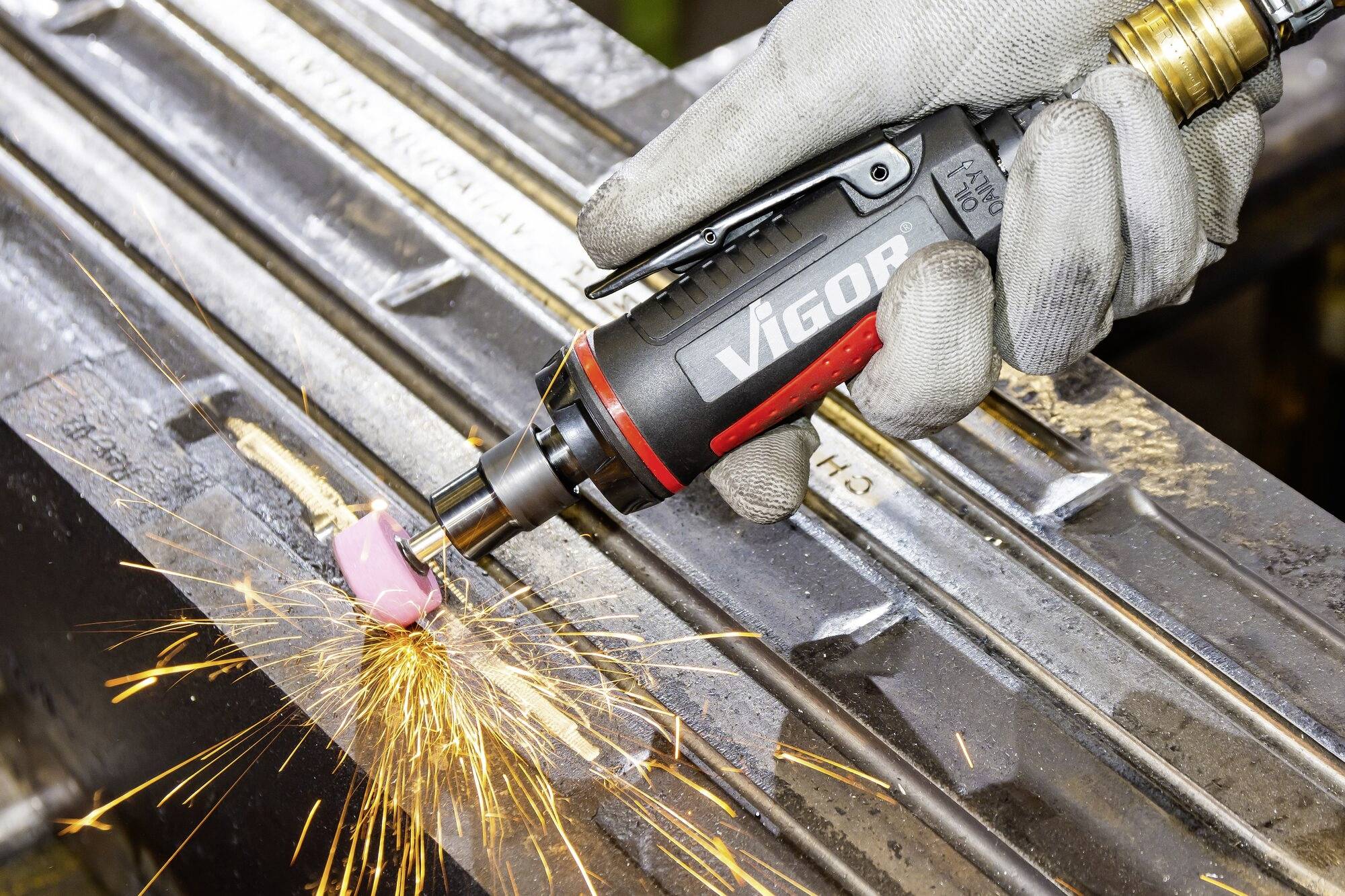 Jedna ręka trzyma szlifierkę kątową, która rozpryskuje iskry podczas obrabiania metalu. Na narzędziu widoczna jest nazwa marki 'VIGOR'.