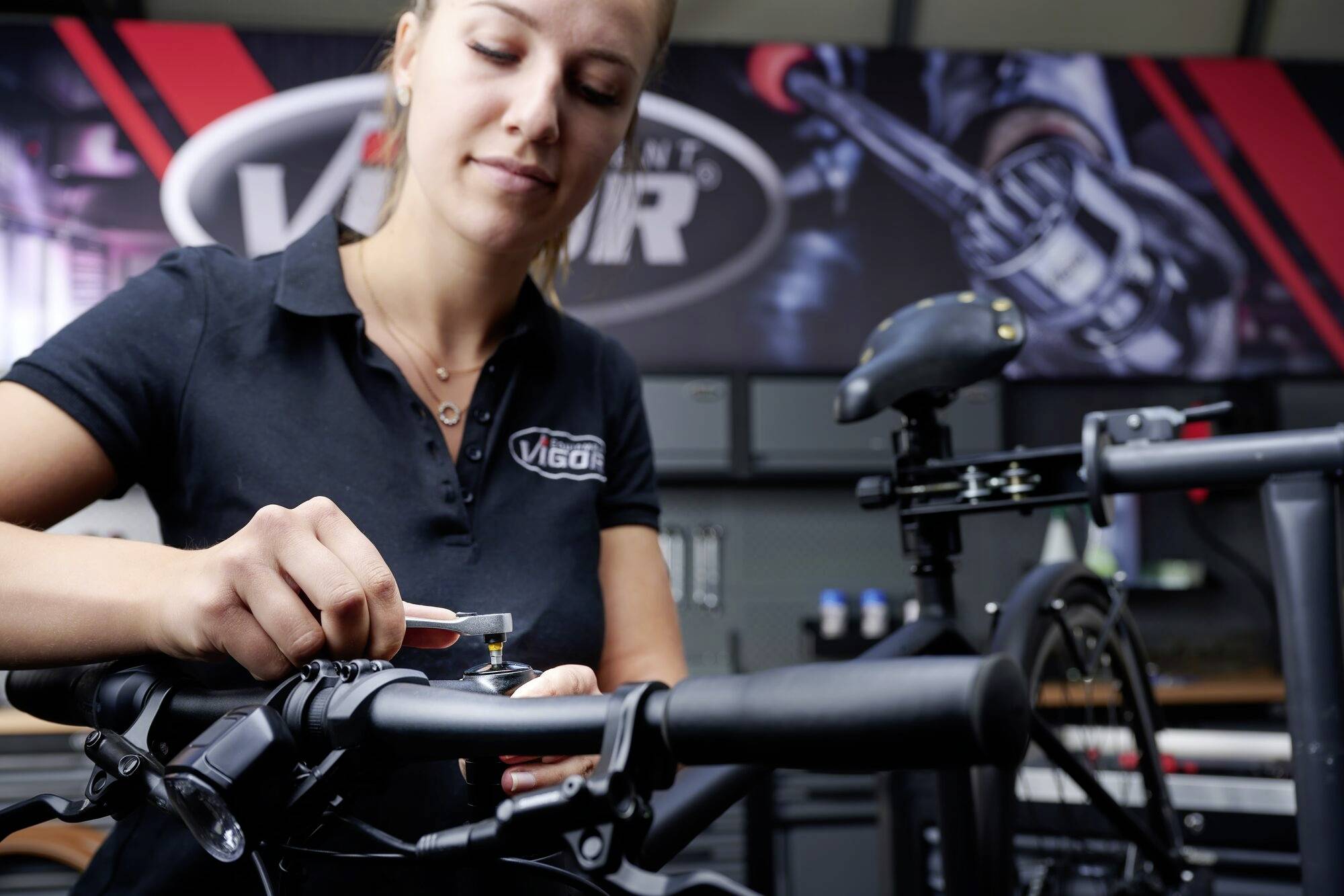 Kobieta pracuje przy kierownicy roweru w warsztacie. W tle widać duży szyld z logo 'Vigor'.