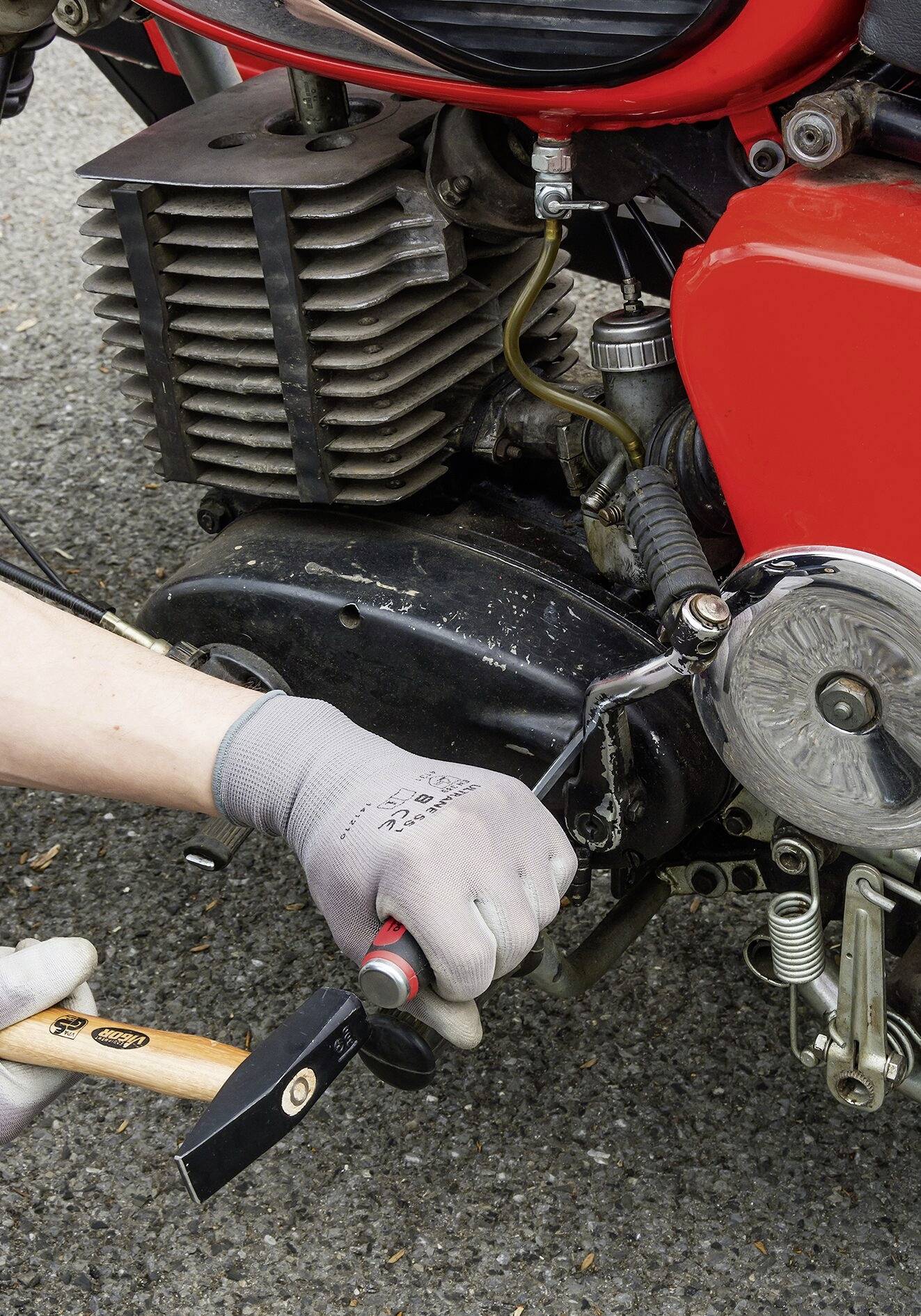 Jedna osoba w rękawicach ochronnych naprawia silnik czerwonego motocykla młotkiem na asfaltowanej drodze.