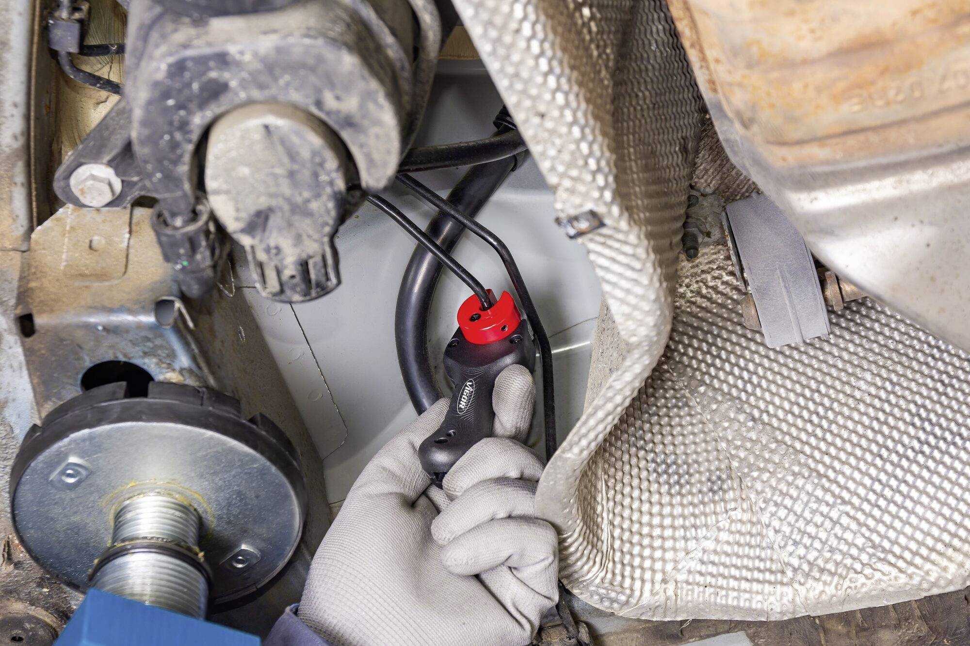 Dłoń w rękawicy trzyma czerwone urządzenie diagnostyczne przy elemencie pojazdu w pobliżu podwozia i układu wydechowego.