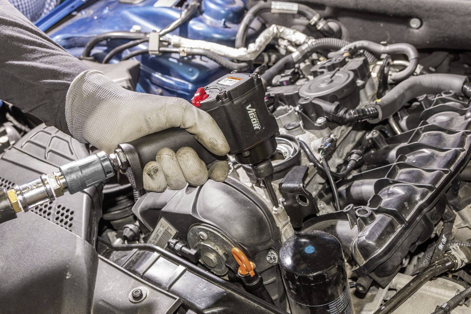 Osoba w rękawicach używa narzędzia pneumatycznego do pracy przy silniku samochodowym. Widoczne są różne komponenty silnika.