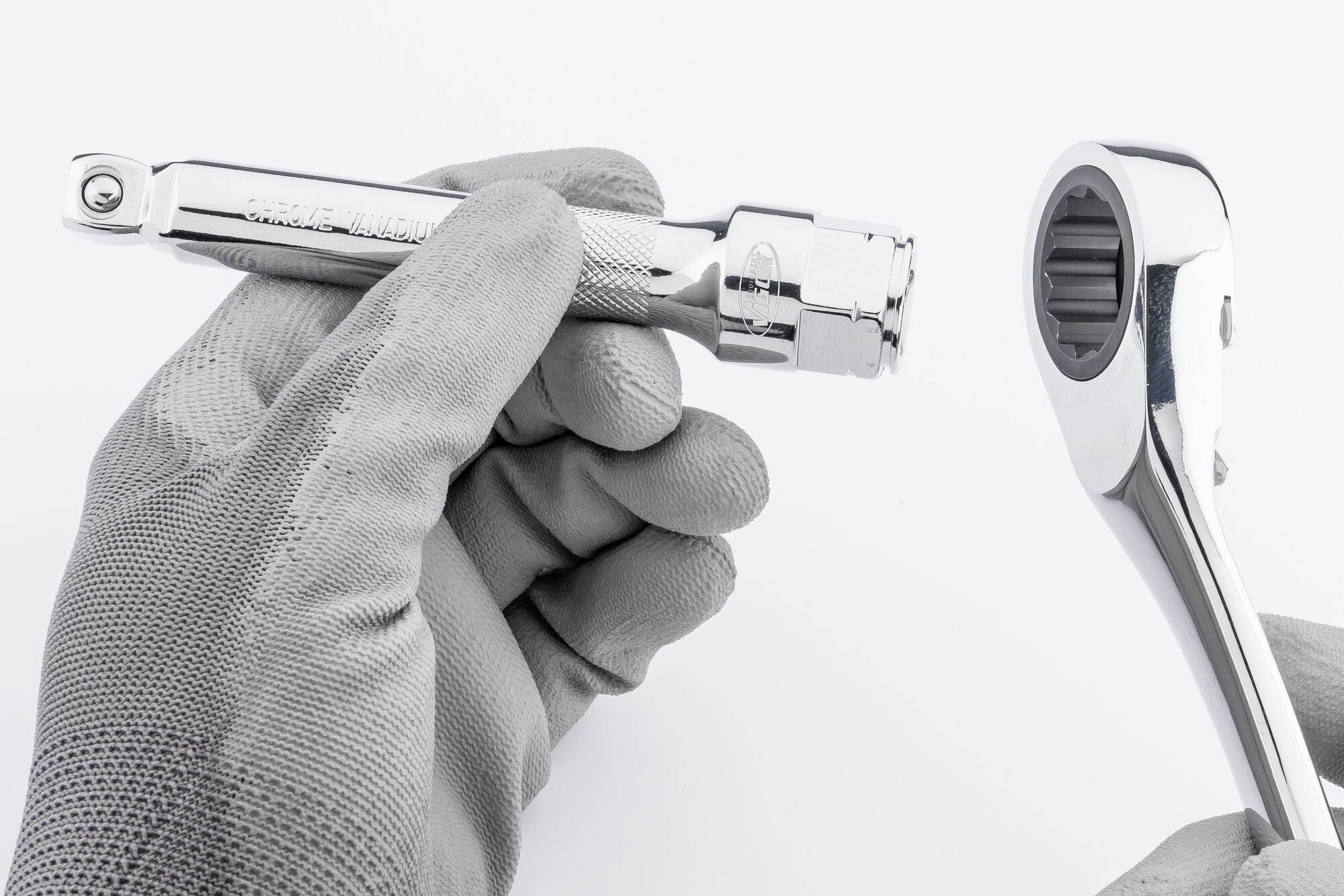 Dłoń w rękawicy roboczej trzyma klucz nasadowy, podczas gdy obok leży klucz zapadkowy. Oba narzędzia są srebrnego koloru.