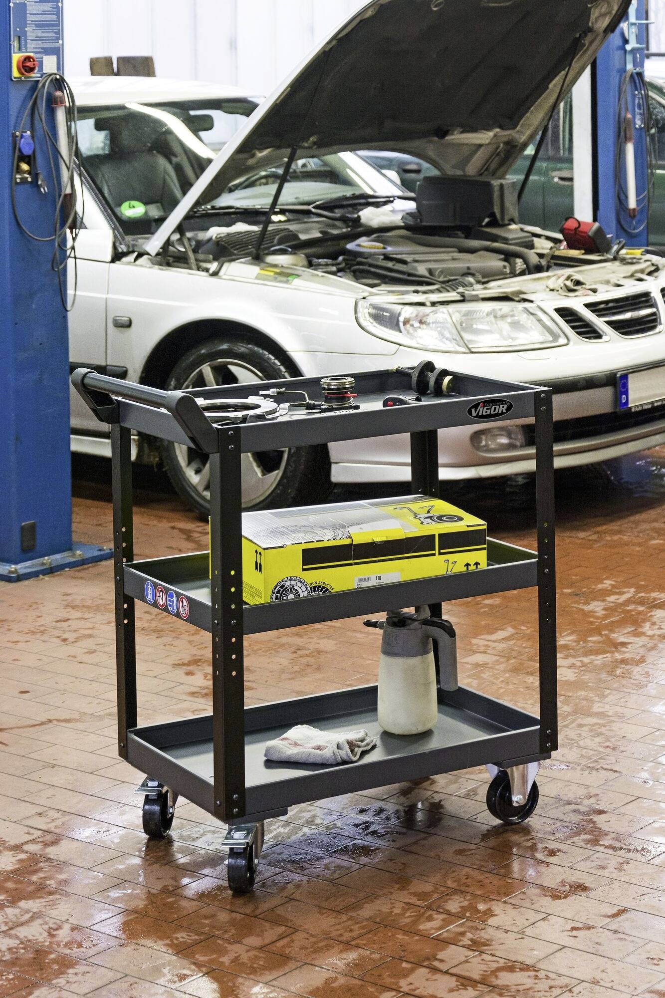 Wózek narzędziowy w warsztacie samochodowym, w tle samochód z otwartą maską silnika zawieszony na podnośniku.