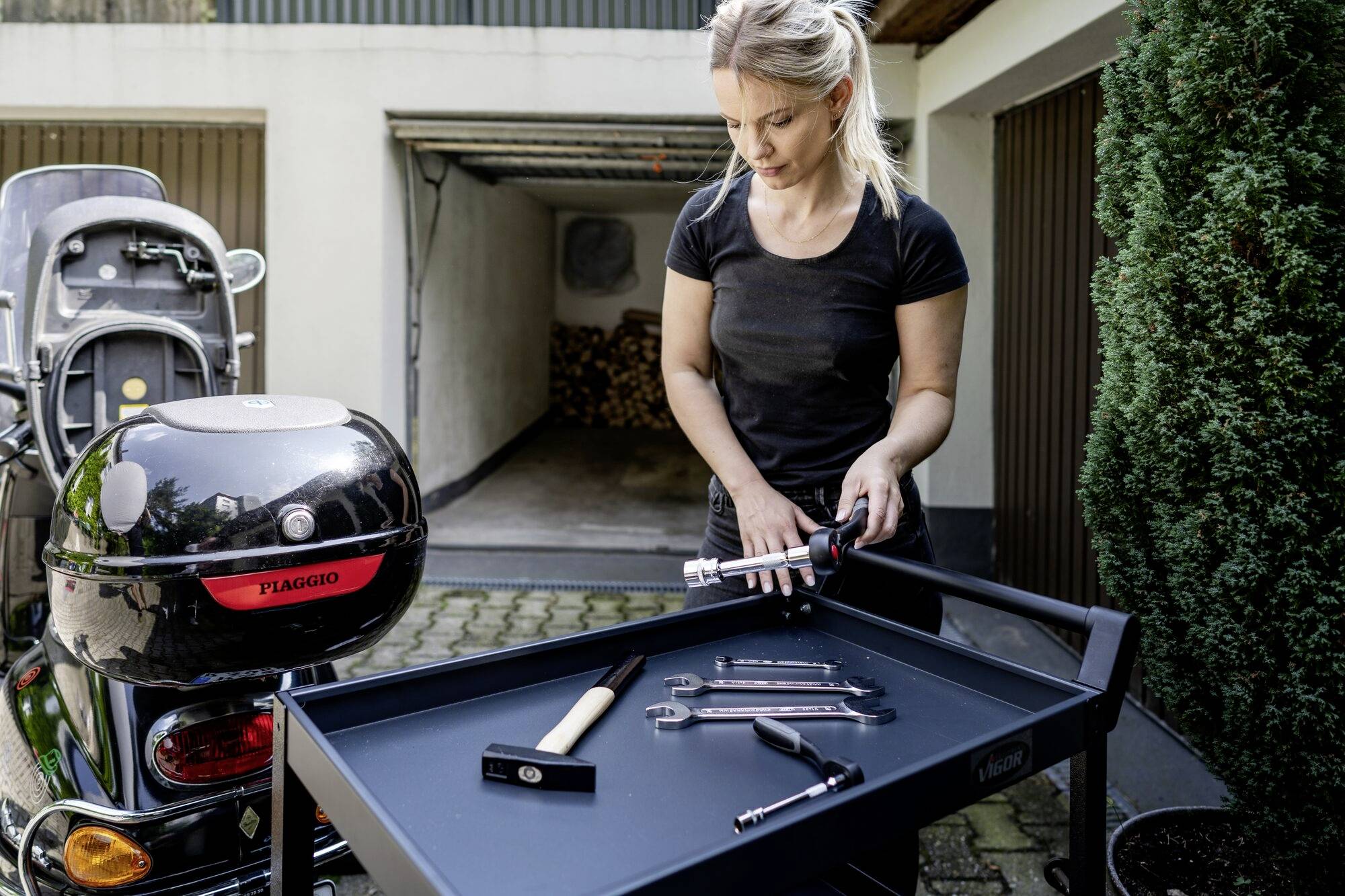 Osoba ustawia czarny wózek narzędziowy z różnymi narzędziami na wolnym powietrzu. W tle stoi motocykl i otwarta garaż.