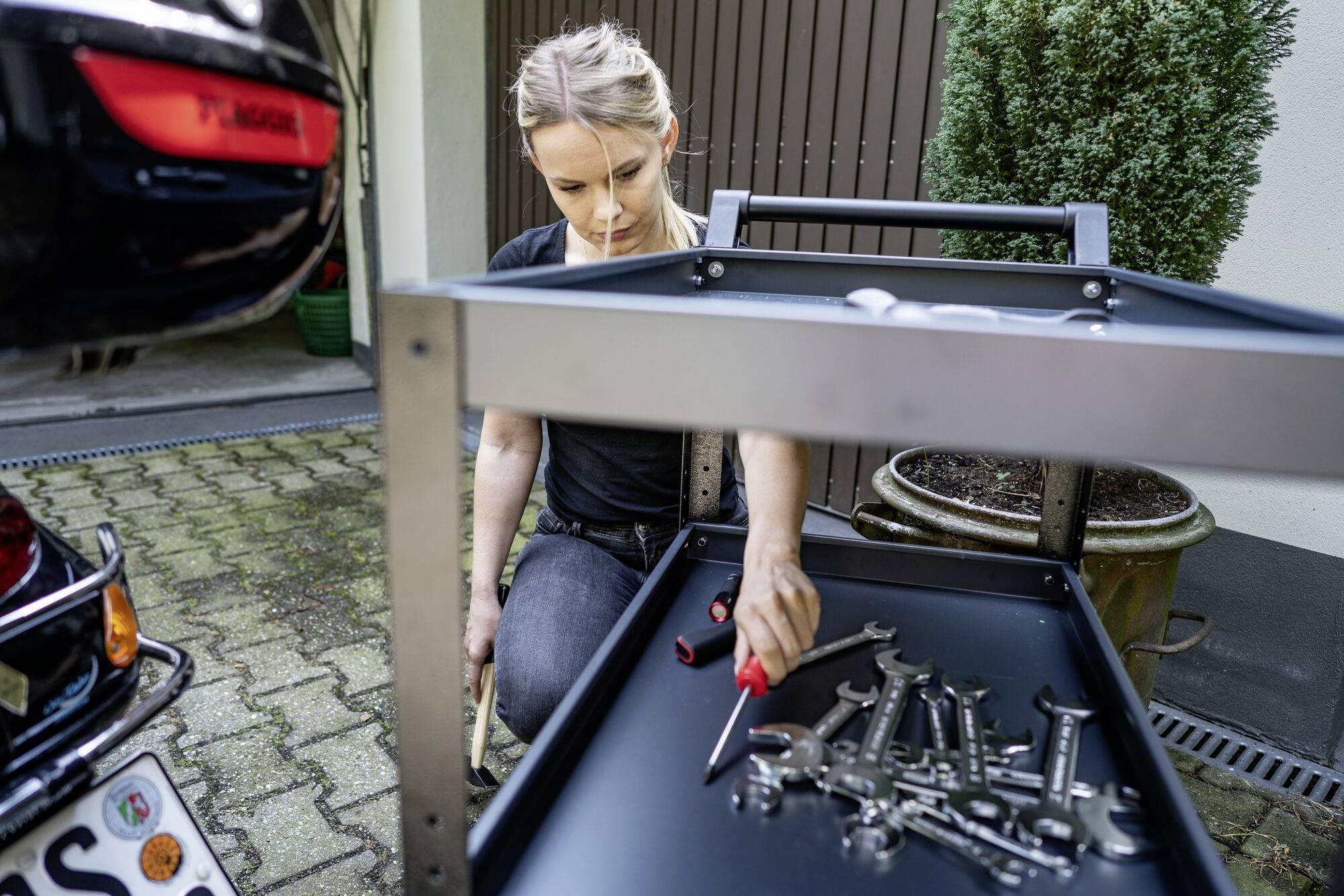 Osoba organizuje narzędzia na wózku warsztatowym obok samochodu w garażu. Trzyma w ręku śrubokręt.