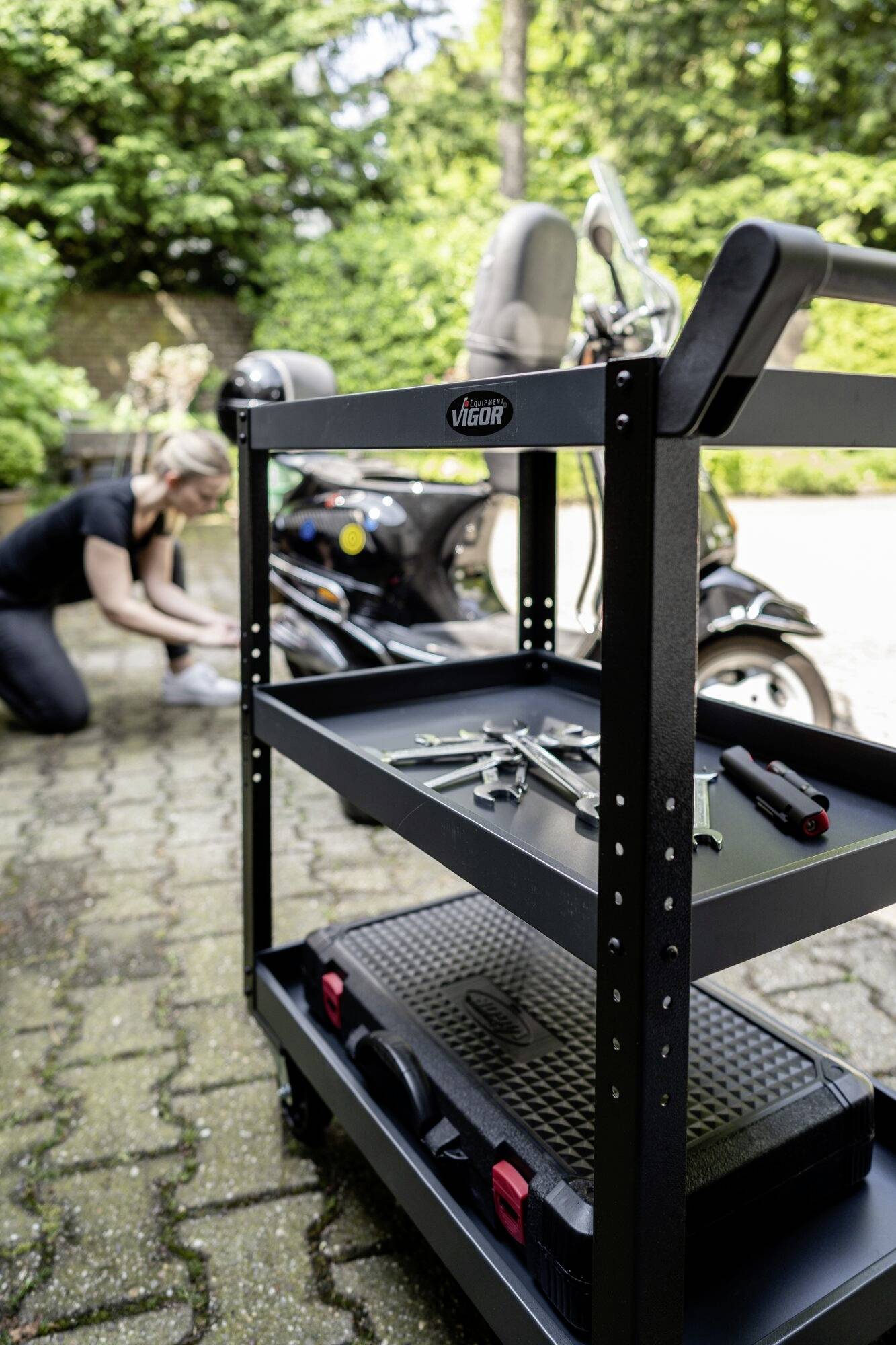 Wózek warsztatowy z narzędziami stoi na pierwszym planie, podczas gdy w tle jedna osoba pracuje przy skuterze.