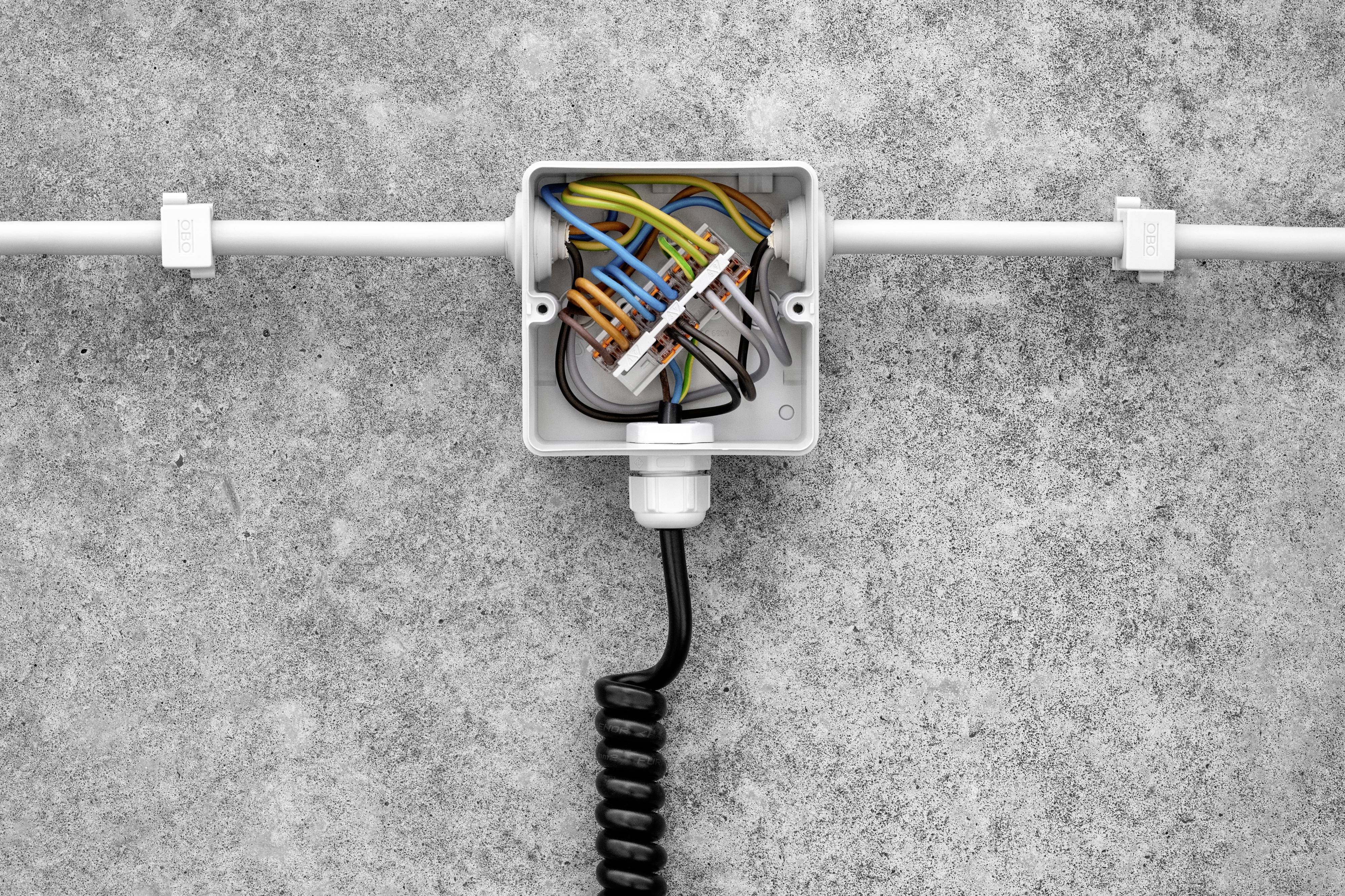 Otwarty rozdzielacz elektryczny z kolorowymi kablami, zamontowany na szarej ścianie. Czarny kabel wyprowadzony jest w dół.