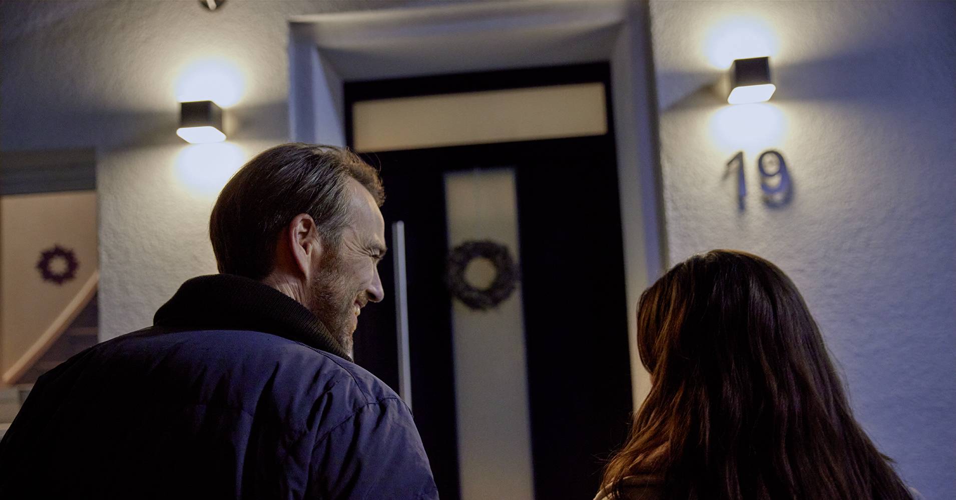 Mężczyzna i kobieta stoją wieczorem przed czarno-białymi drzwiami wejściowymi, ozdobionymi wieńcem, z widocznym numerem domu 19.