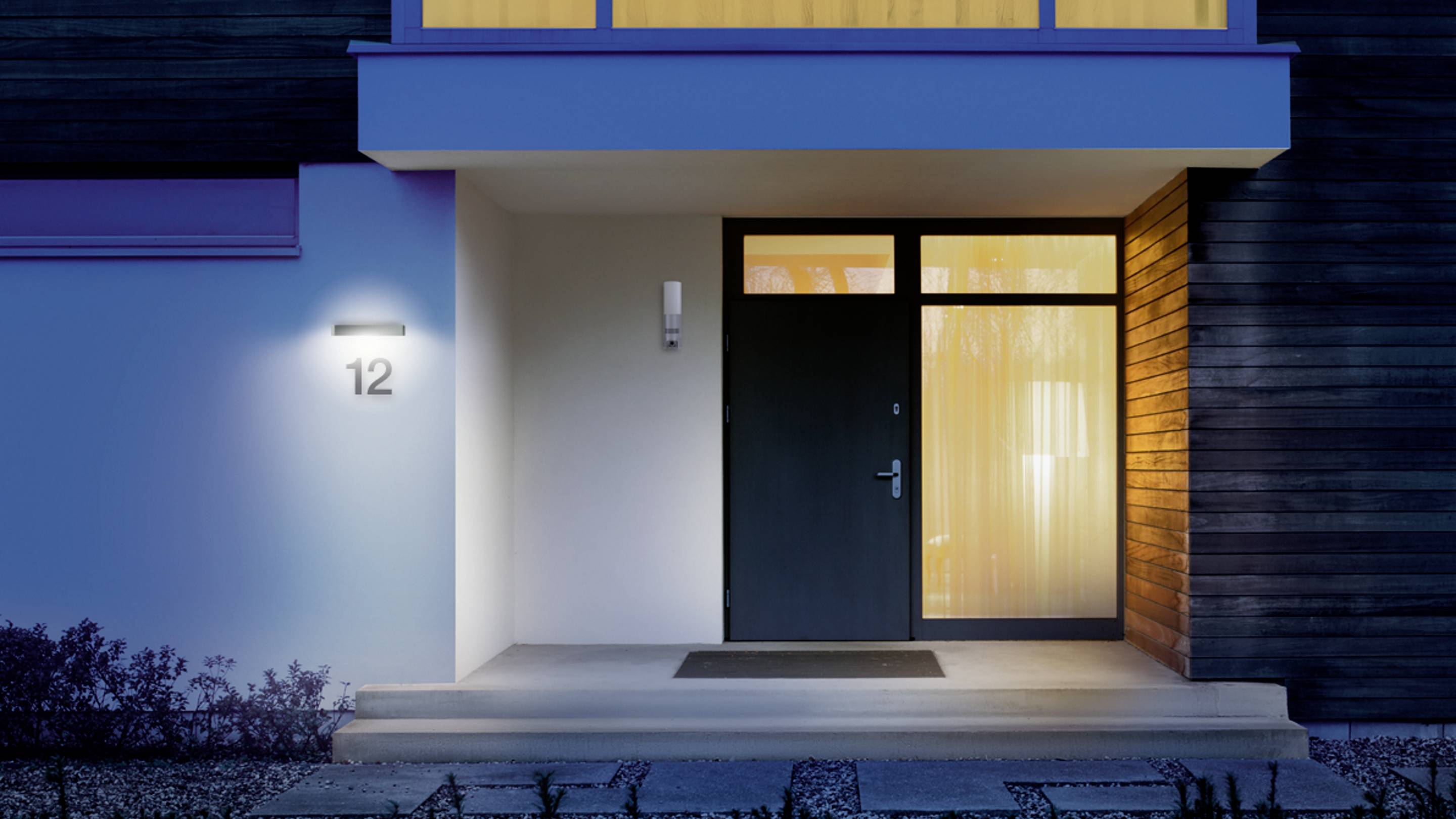 Wejście do nowoczesnego domu nocą, z podświetlonym numerem domu '12' na białej ścianie i oświetlonym oknem za ciemnymi drzwiami.