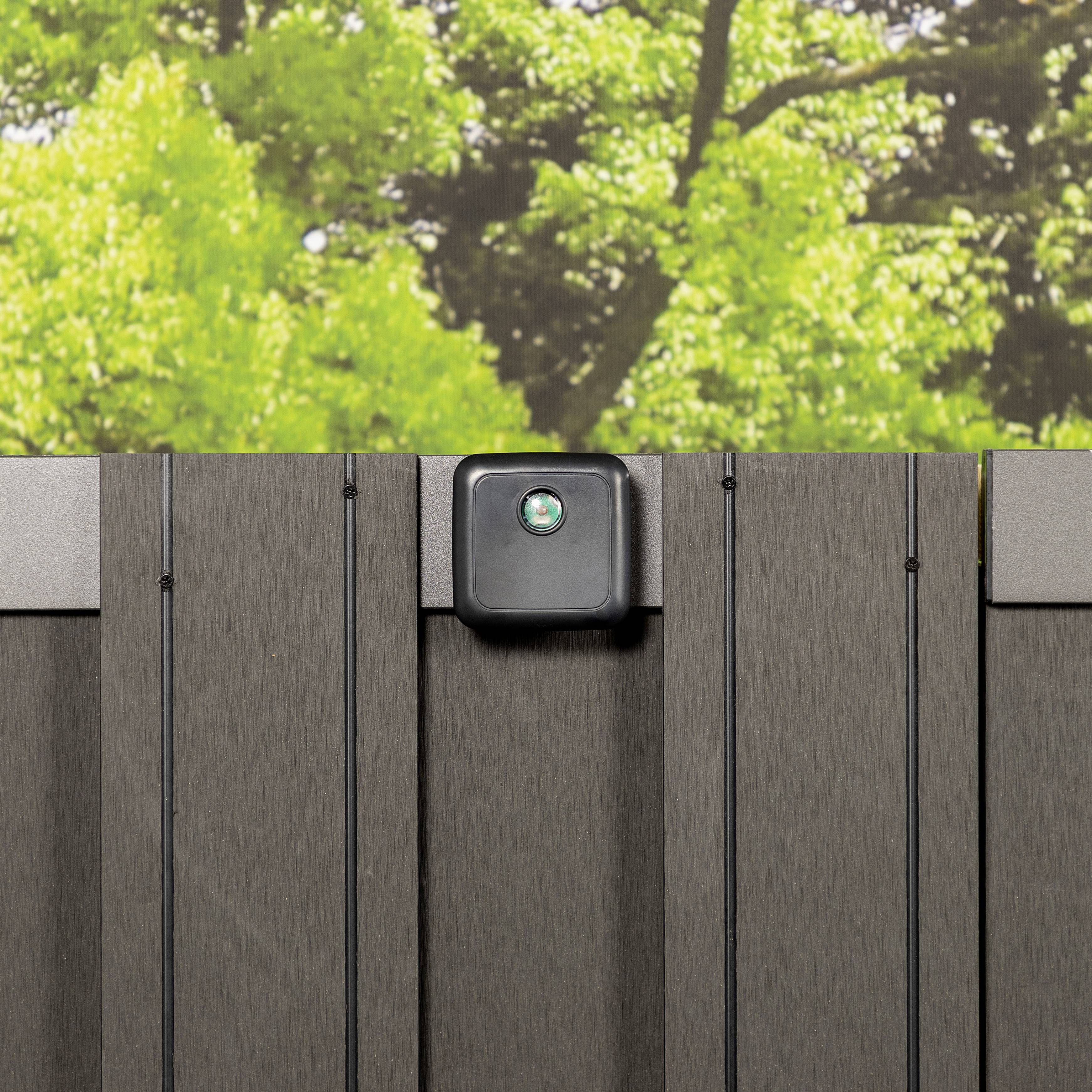 Czarne urządzenie zewnętrzne, prawdopodobnie kamera lub światło, jest zamocowane na pionowej, szarej płycie ogrodzenia; w tle widoczne są drzewa.