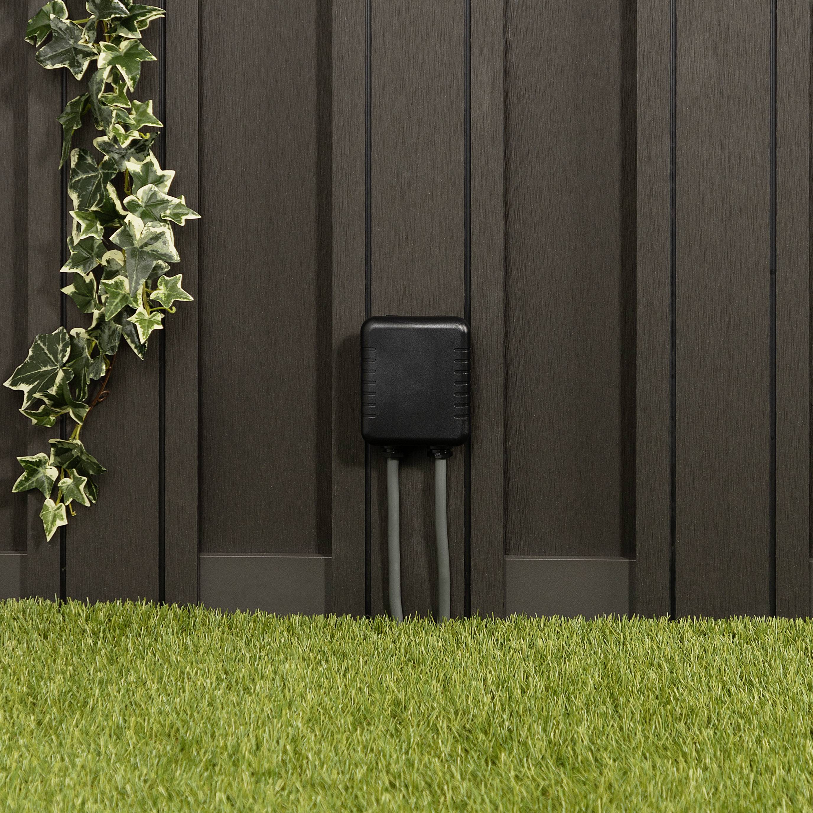Elektryczne urządzenie z trzema przewodami jest zamocowane na nowoczesnej ścianie. Po lewej stronie obok niego zwisa roślina pnąca. Przed nią rozciąga się zielony trawnik.