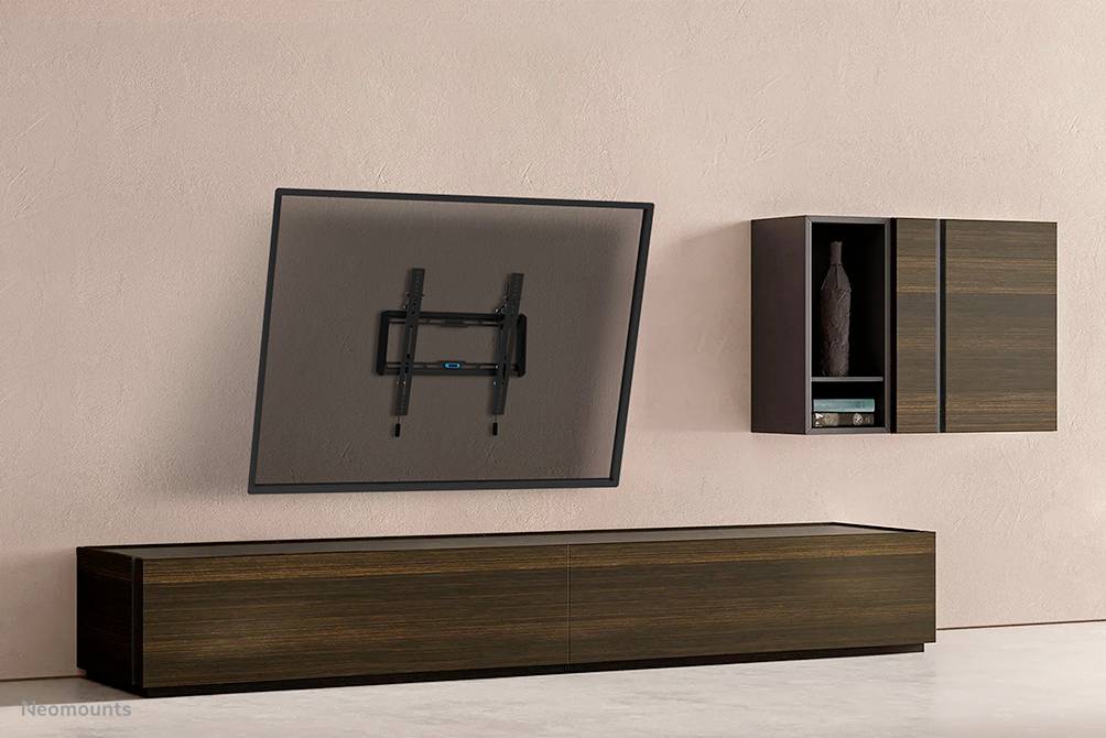 Telewizor na regulowanym uchwycie ściennym obok drewnianej szafki i regału na tle różowej ściany. Nowoczesne wnętrze o minimalistycznym designie.