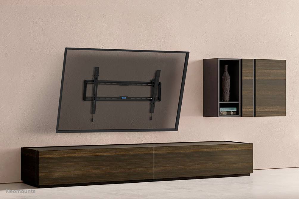 Uchwyt ścienny do telewizora przedstawia nachylony telewizor. Pod uchwytem stoi drewniana szafka. Po prawej stronie na ścianie wisi szafka.