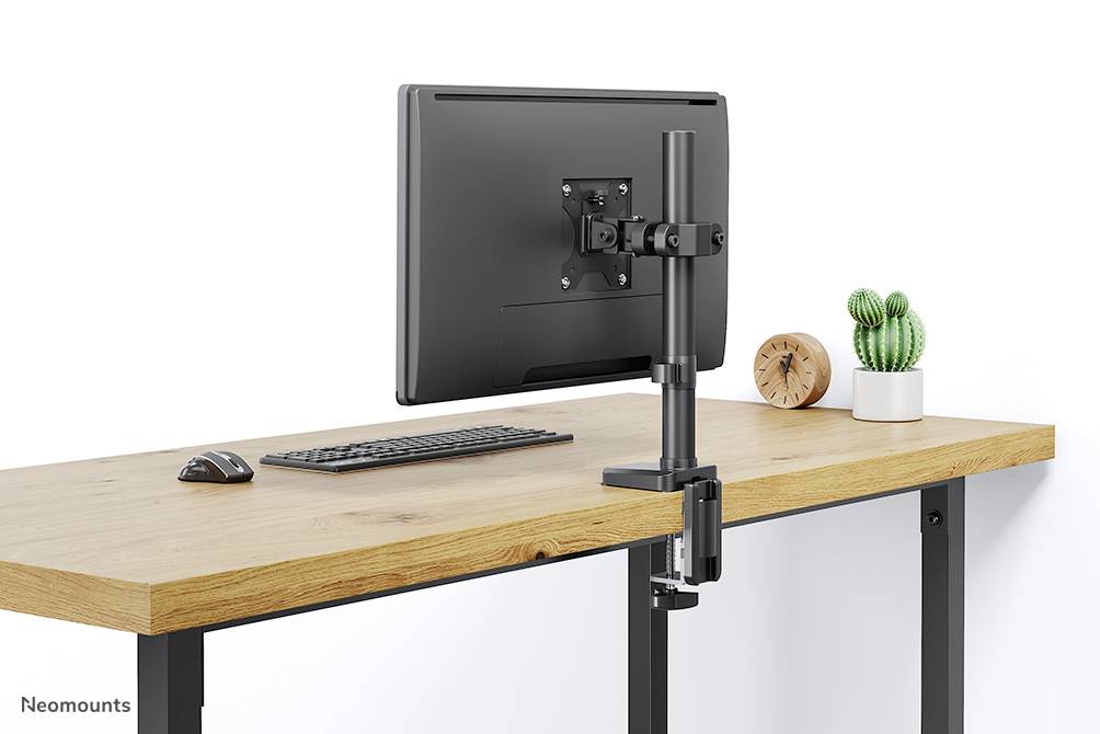 Uchwyt na monitor na drewnianym stole, obok klawiatury i myszy. W tle roślina i zegar.