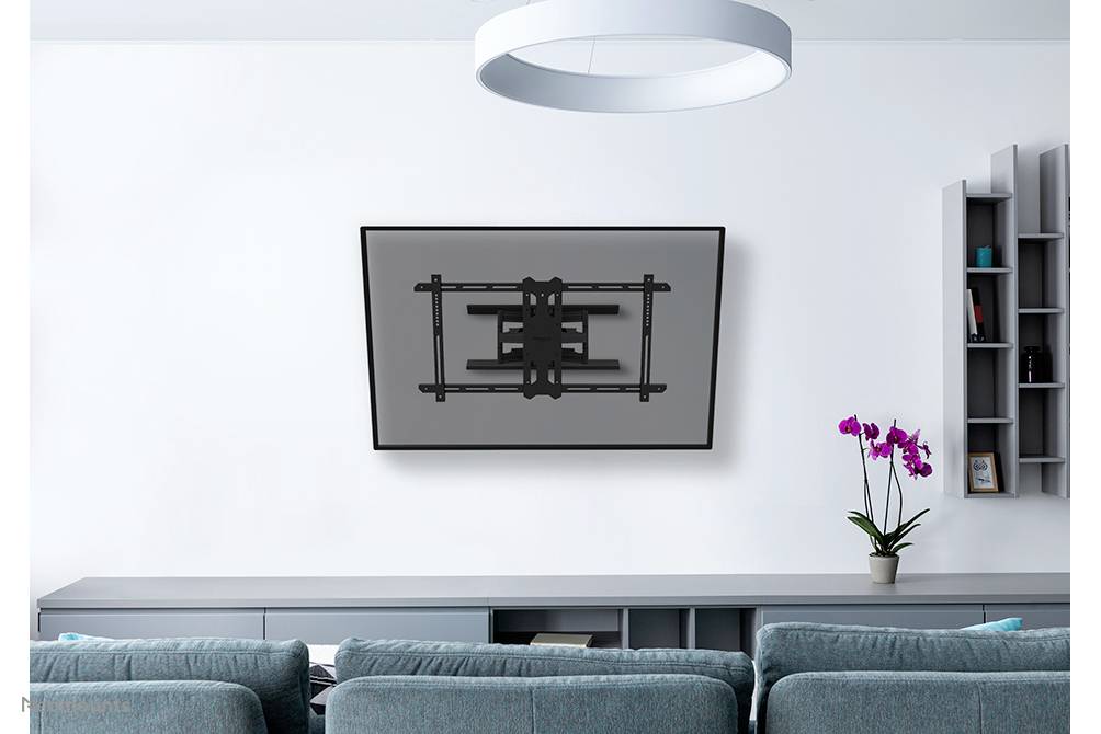 Płaski telewizor wisi na ścianie w nowoczesnym salonie. Na pierwszym planie znajduje się szara sofa, obok której stoi orchidea.