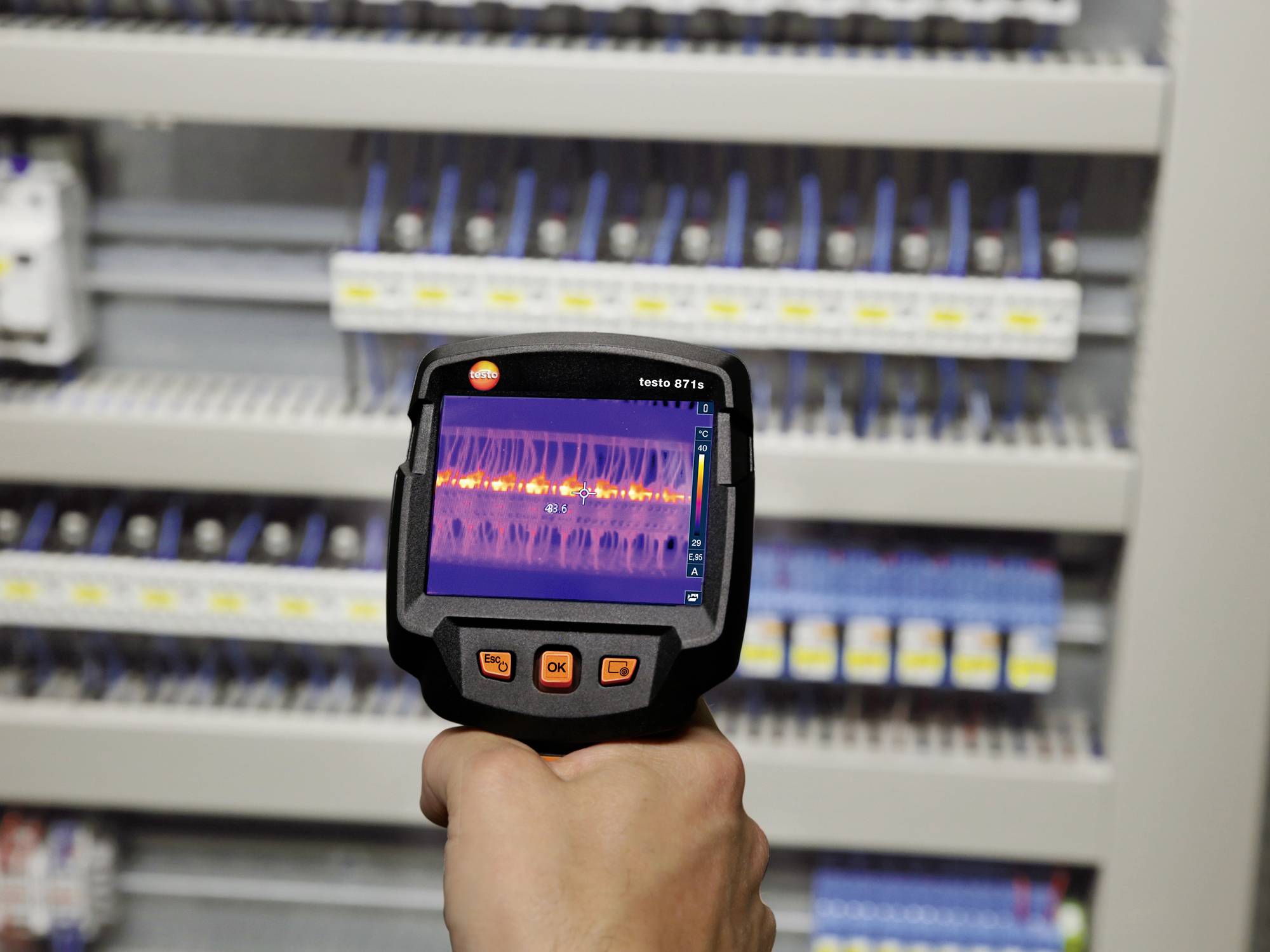 Dłoń trzyma kamerę termowizyjną, która skanuje skrzynkę bezpieczników. Wyświetlacz pokazuje gradacje kolorów, wskazujące na różnice temperatury.