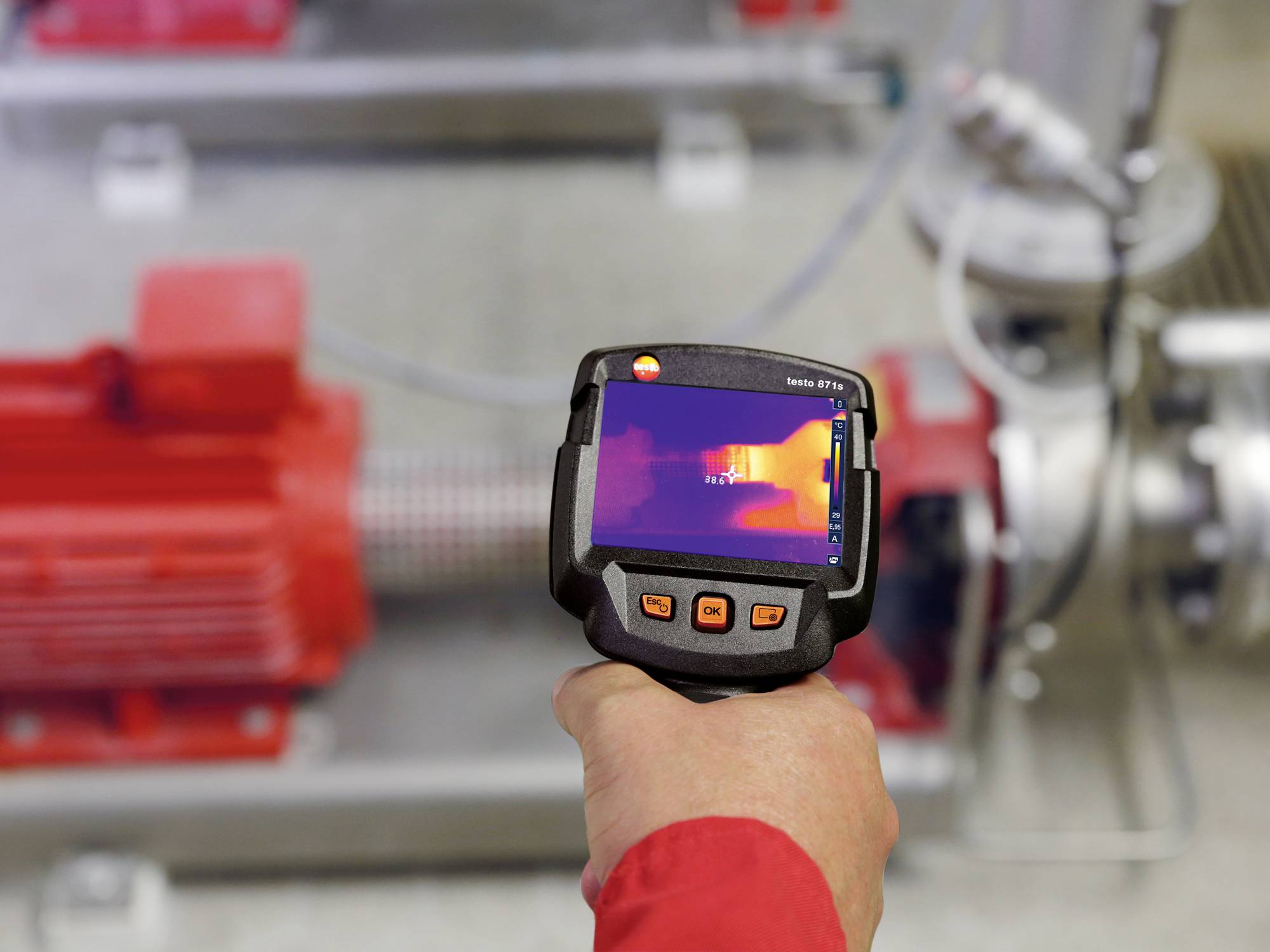 Osoba trzyma kamerę termowizyjną i kieruje ją na czerwoną maszynę, aby zmierzyć jej temperaturę. Ekran kamery termicznej jest widoczny.