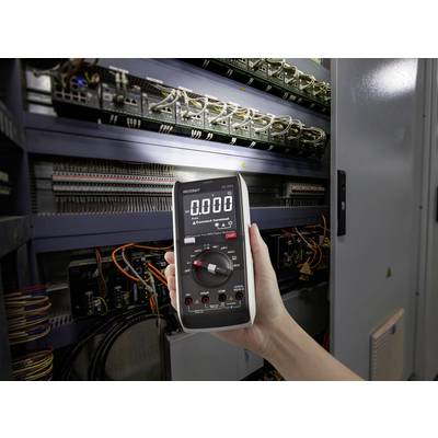 Jedna ręka trzyma multimetr cyfrowy ustawiony na 0,000 przed otwartą szafką sterowniczą pełną kabli i elementów elektronicznych.