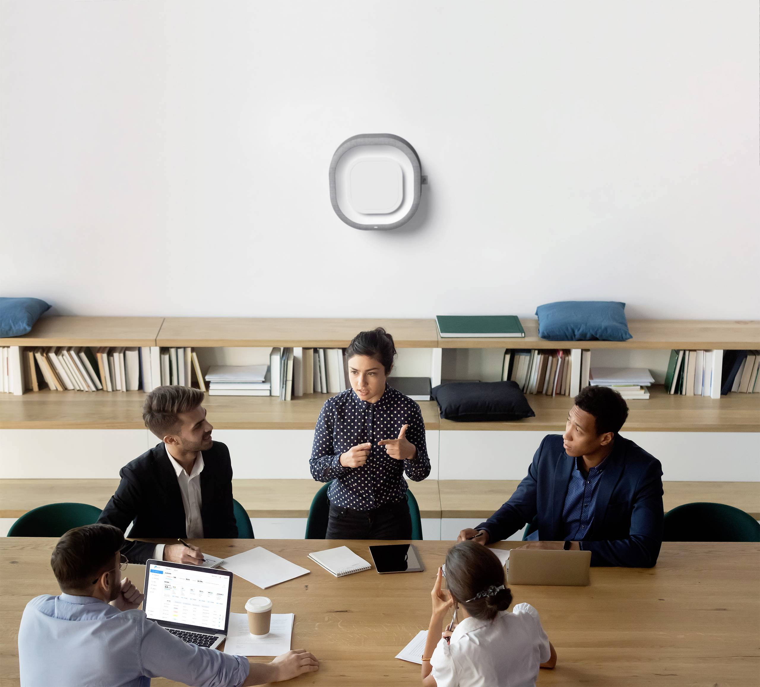 Pięć osób siedzi w sali konferencyjnej, podczas gdy kobieta stojąca pośrodku prowadzi dyskusję. W tle widać regał wypełniony książkami.