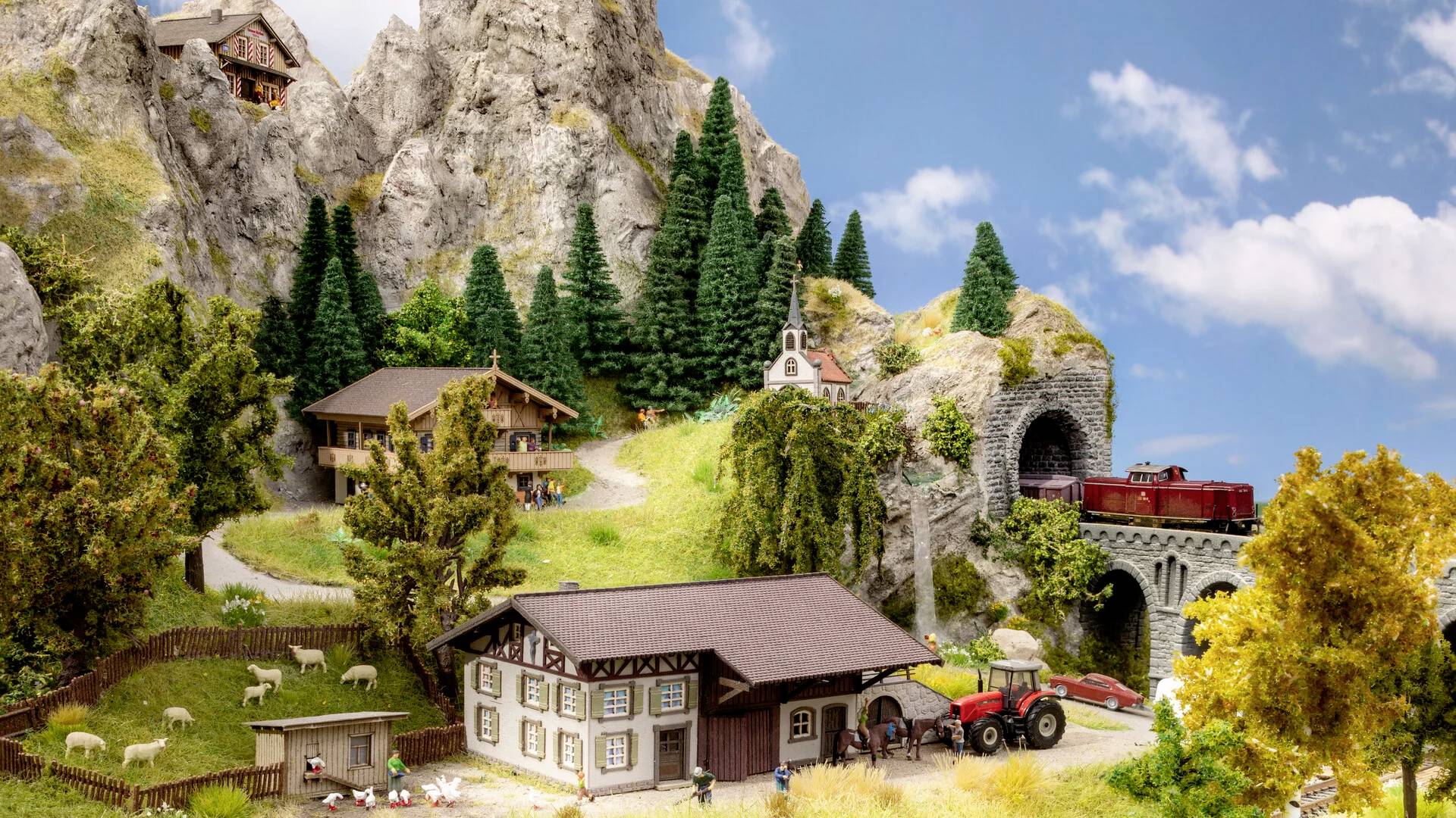 Miniaturowy model alpejskiego krajobrazu z górami, świerkami, domami wiejskimi, ciągnikiem i pociągiem w tunelu. Spokojna scena wiejska.
