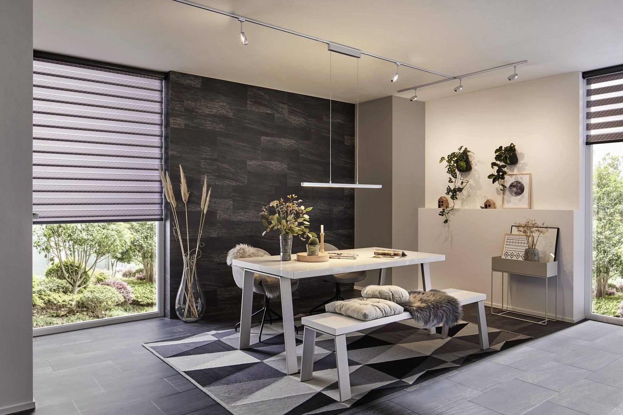 Jadalnia z nowoczesnym stołem, dwiema ławkami i wiszącą lampą. Ciemna ściana, rośliny i dekoracje tworzą przytulną atmosferę.