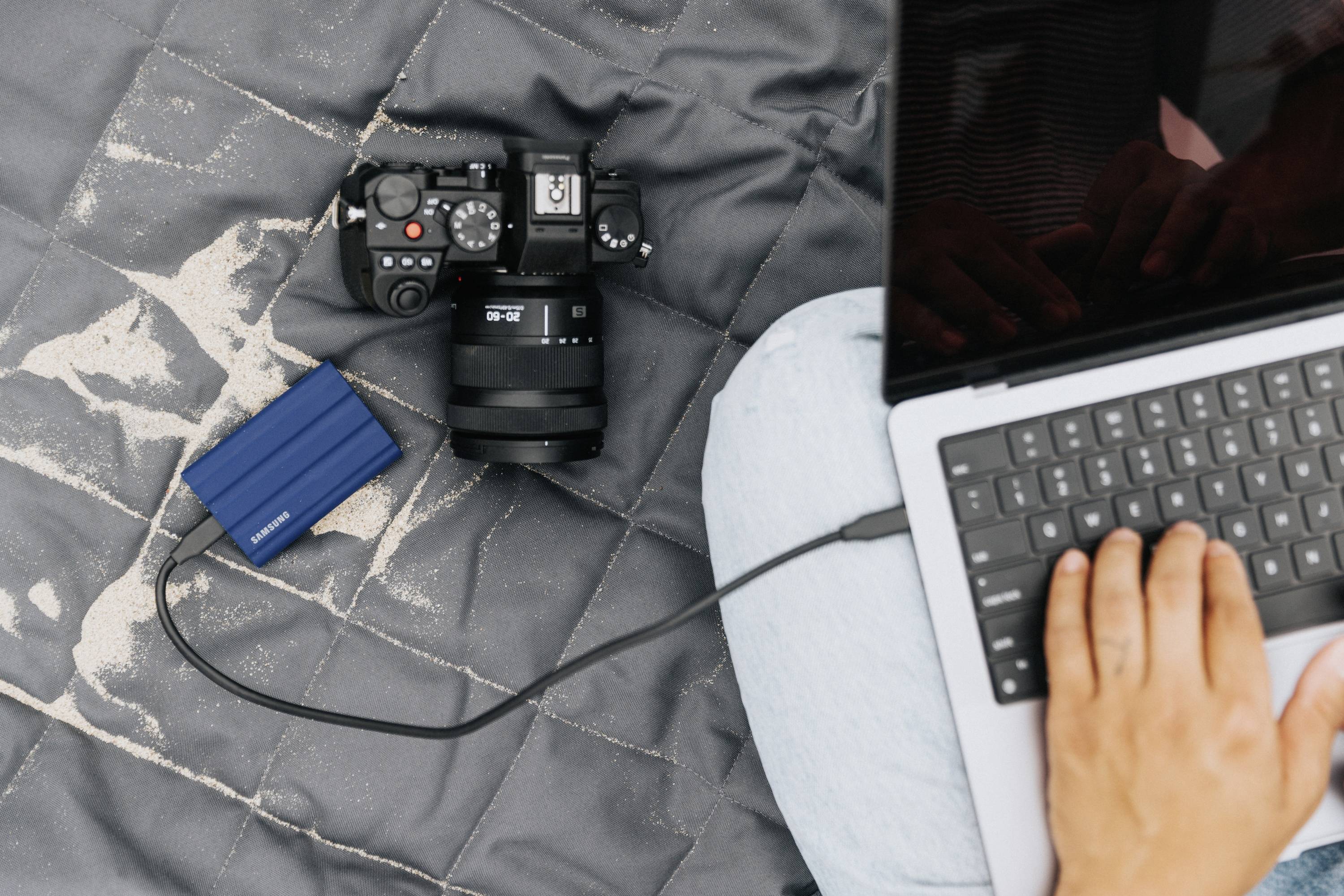 Osoba pracuje na laptopie. Obok laptopa, na kocu, leżą kamera i zewnętrzny dysk twardy.