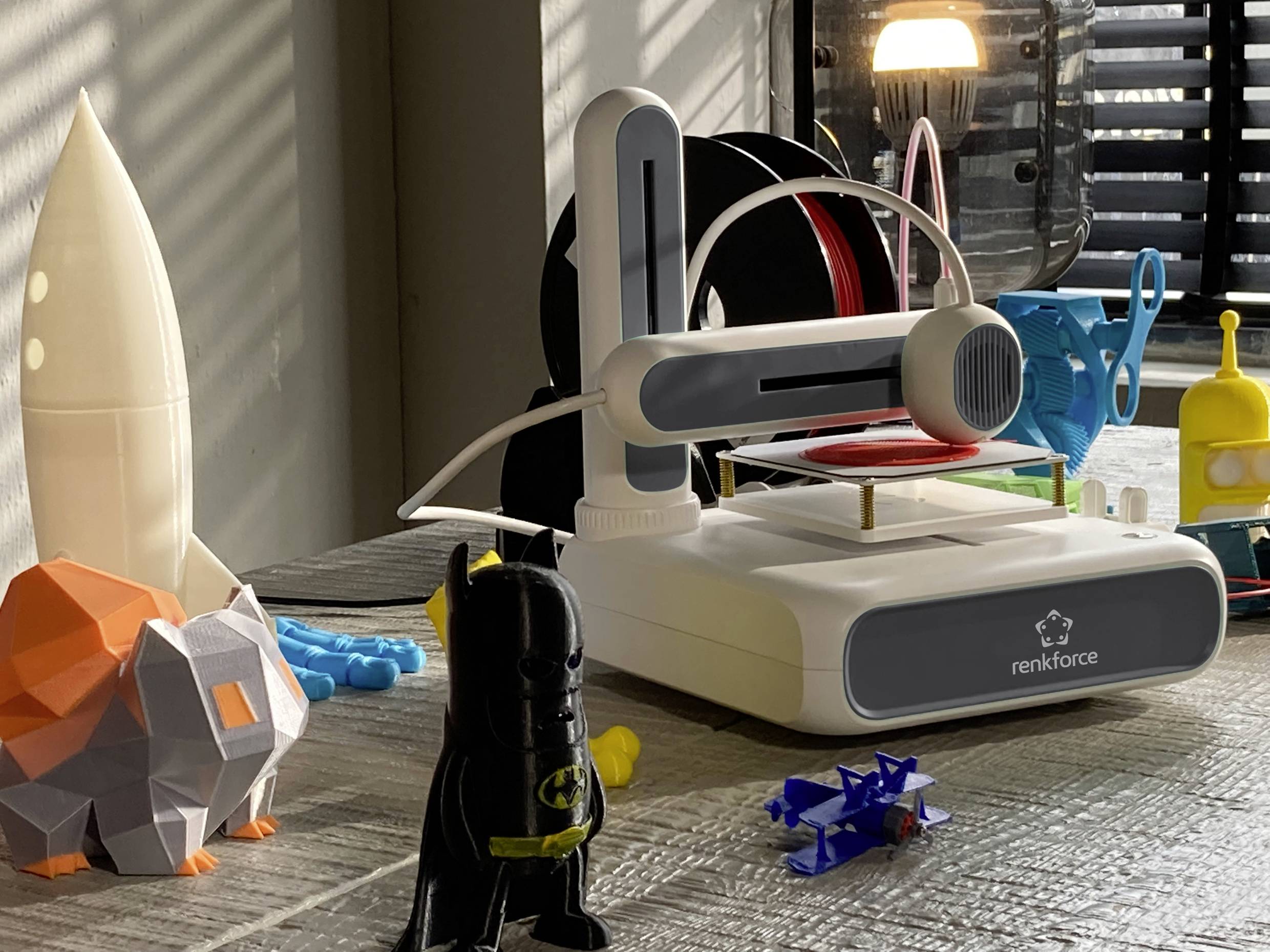 Drukarka 3D z logo 'renkforce' stoi na stole, otoczona kolorowymi figurkami wydrukowanymi w technologii 3D, takimi jak rakieta i postać z kreskówki.
