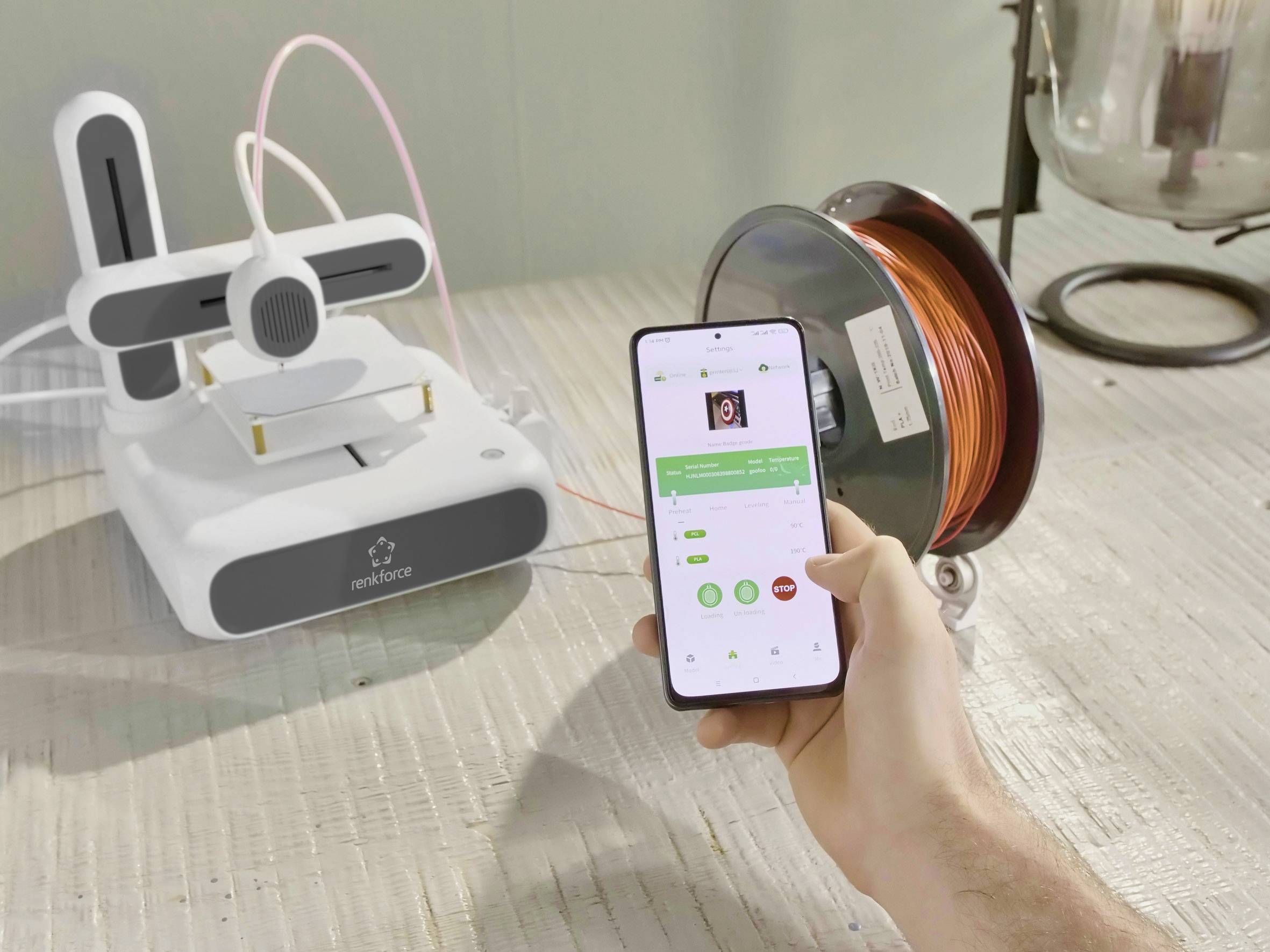 Dłoń trzyma smartfon wyświetlający aplikację sterującą drukarką 3D. Drukarka umieszczona jest obok szpuli z czerwonym filamentem.