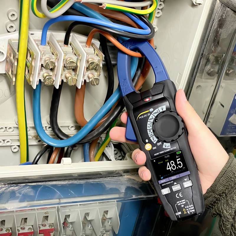 Osoba używa cyfrowego multimetru do pomiaru napięć na tablicy elektrycznej. Widoczne są kolorowe przewody.