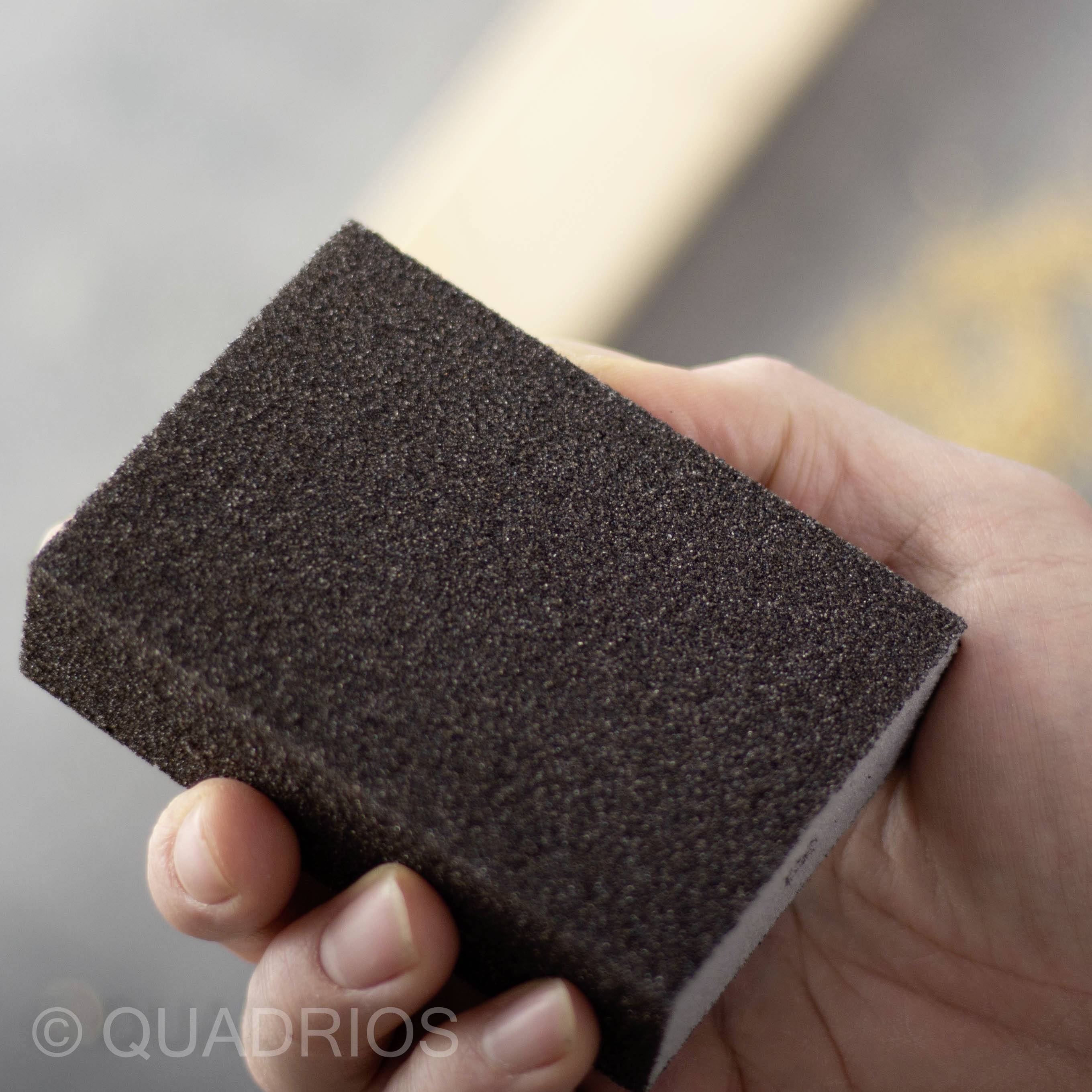 Jedna ręka trzyma czarną gąbkę ścierną, gotową do szlifowania. W tle niewyraźnie widoczne są wióry drewniane.