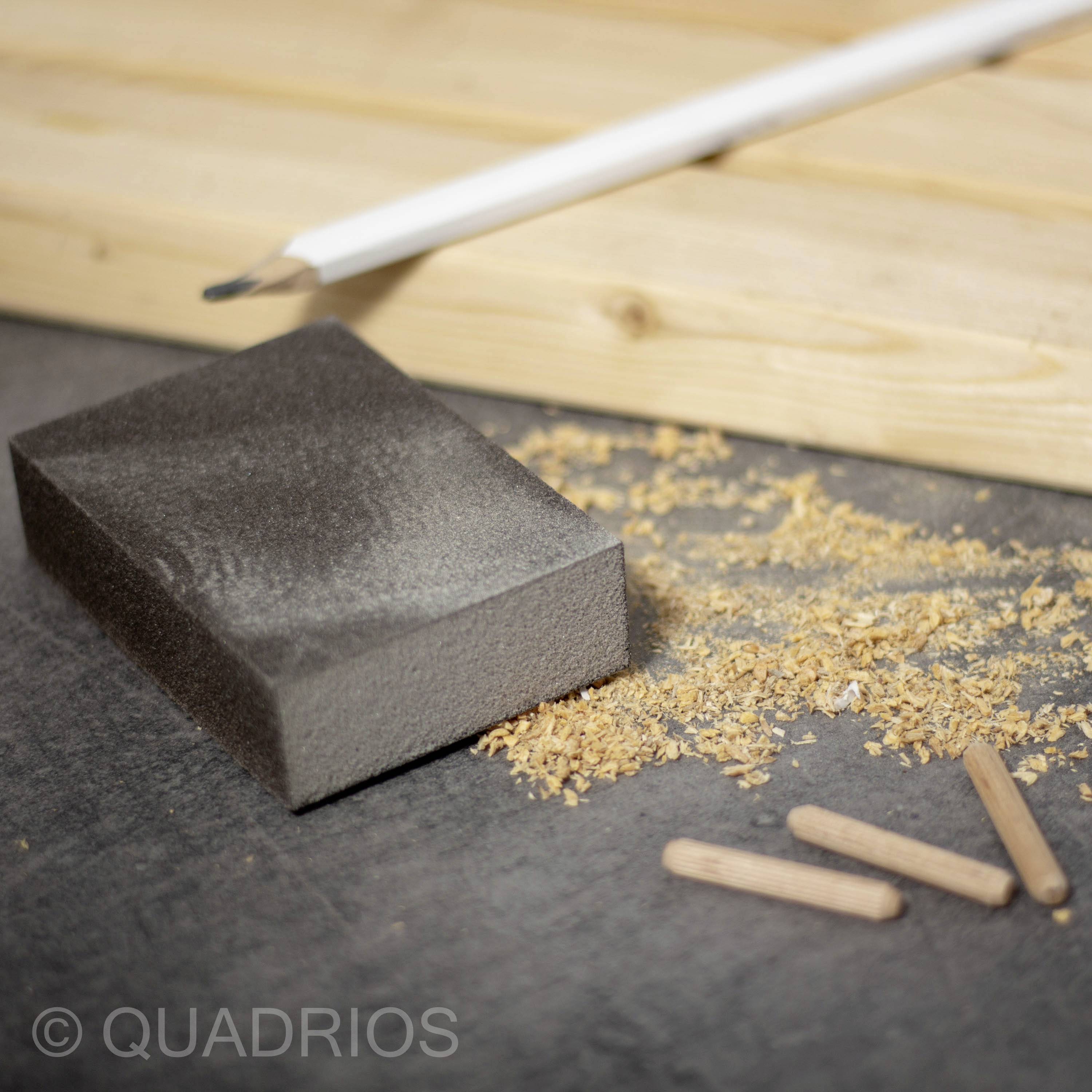 Blok szlifierski i ołówek na drewnianym stole, otoczony trocinami i drewnianymi kołkami. Narzędzia i materiały do projektu stolarskiego.