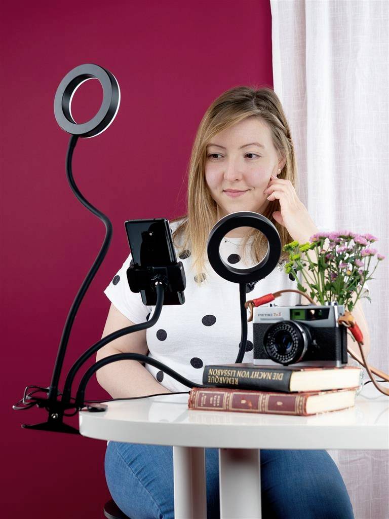 Osoba siedzi przy stole z podstawką do smartfona i lampą pierścieniową, otoczona kwiatami i książkami.