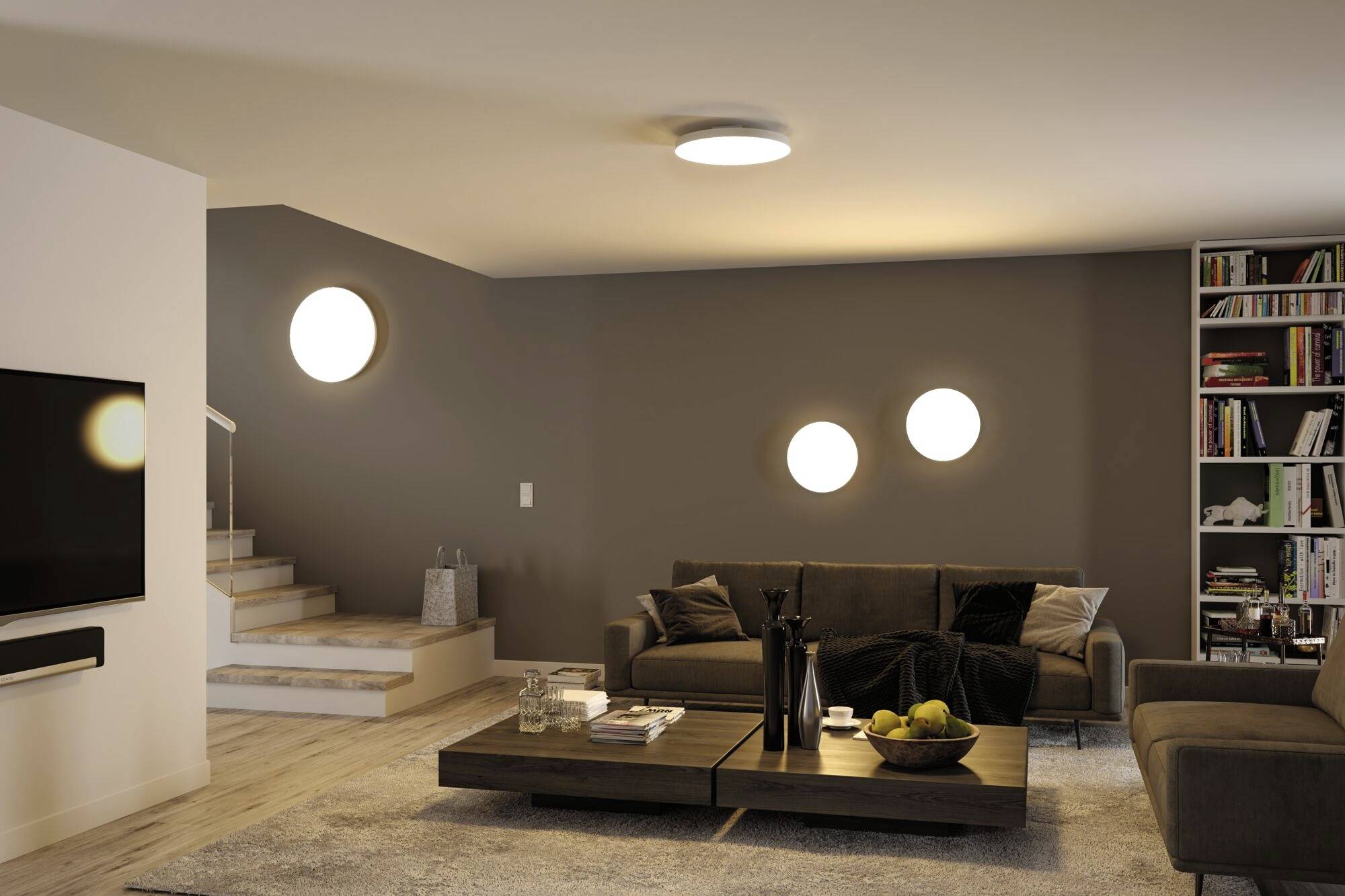 Nowoczesny widok salonu z szarymi ścianami, wygodnymi sofami, regałem na książki i centralnym stolikiem kawowym. Subtelne oświetlenie sufitowe i ścienne.