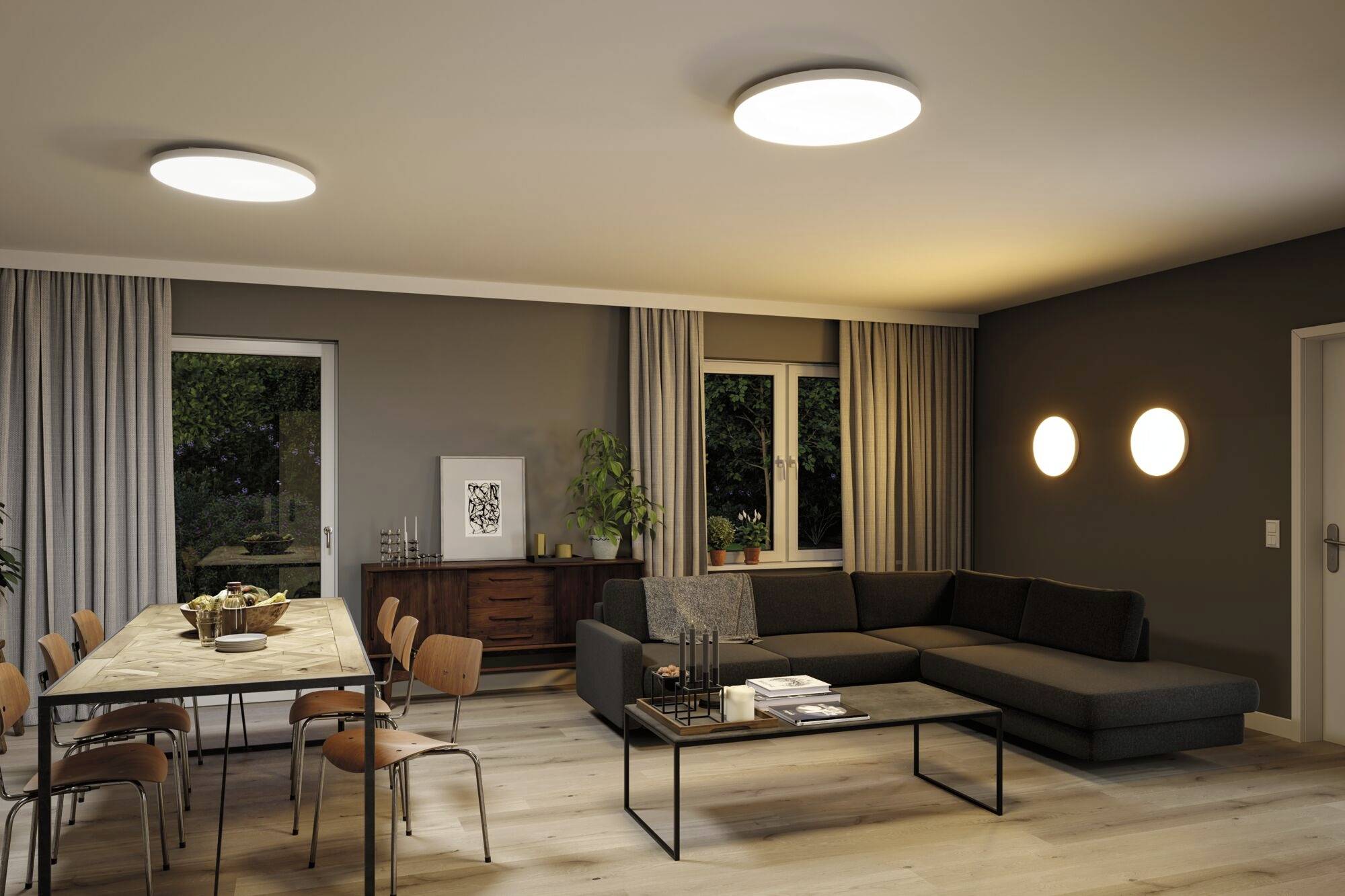 Nowoczesny salon z kanapą, stołem jadalnianym i krzesłami. Dwie lampy sufitowe oświetlają pomieszczenie utrzymane w ciepłych barwach.