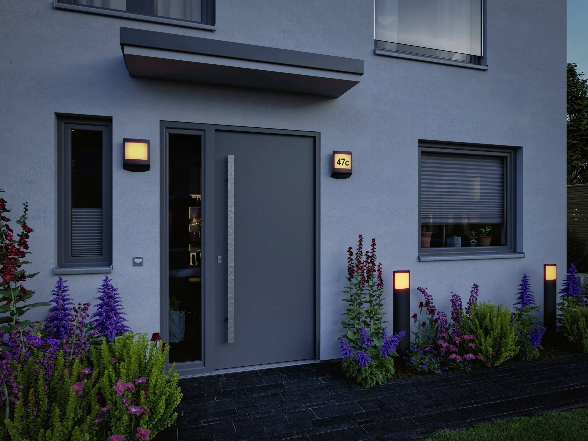 Nowoczesne drzwi wejściowe w ciemnoszarym kolorze na jasnoszarym budynku, otoczone starannie pielęgnowanymi roślinami ogrodowymi i subtelnym oświetleniem zewnętrznym.