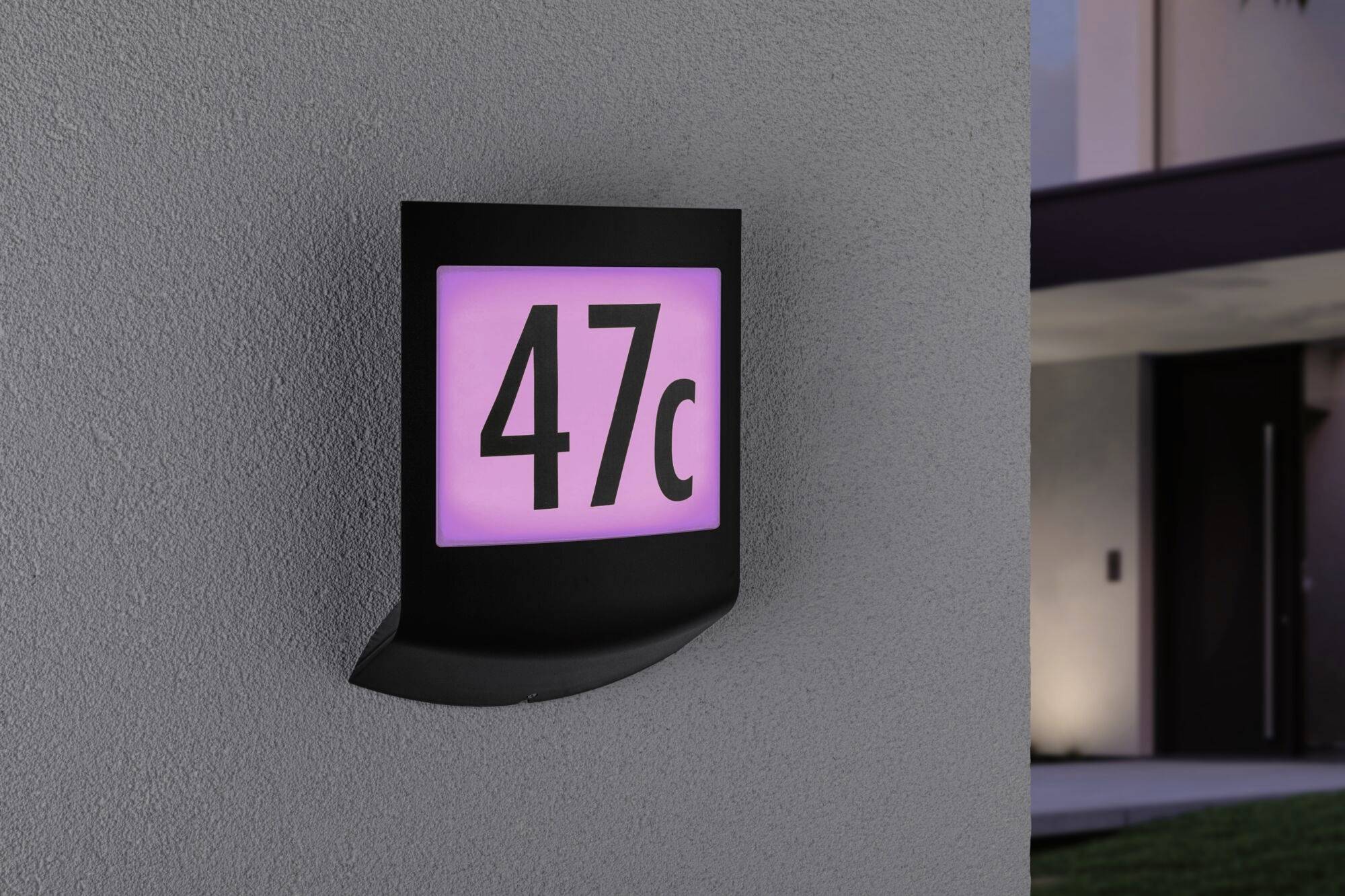 Świecący numerek domu z napisem '47c' zamontowany na szarej ścianie. Otoczenie lekko przyciemnione.