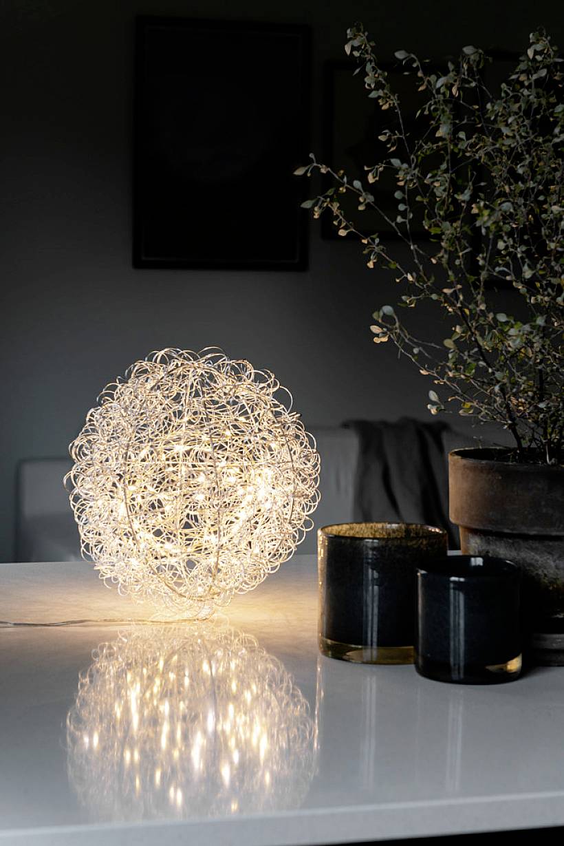 Kula lamp o delikatnym, ażurowym wzorze z drutu ciepło świeci na stole, obok którego stoją dwa czarne świeczniki.