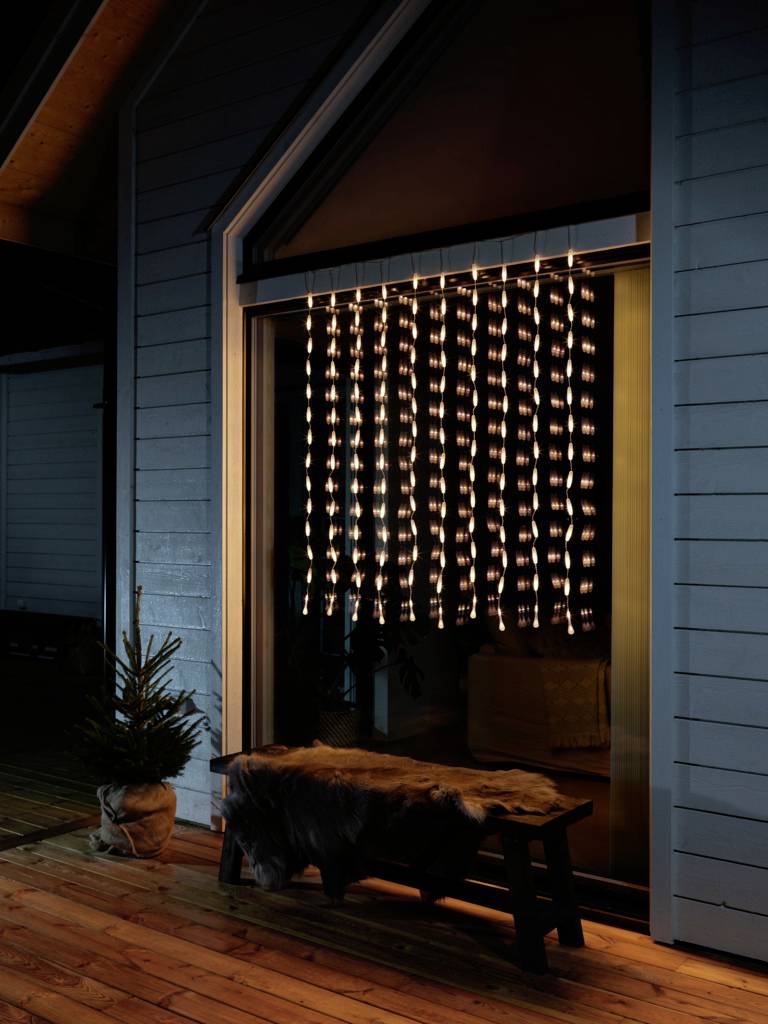 Oświetlona zasłona wisi przed drzwiami tarasowymi nocą; po lewej stronie stoi mała choinka.
