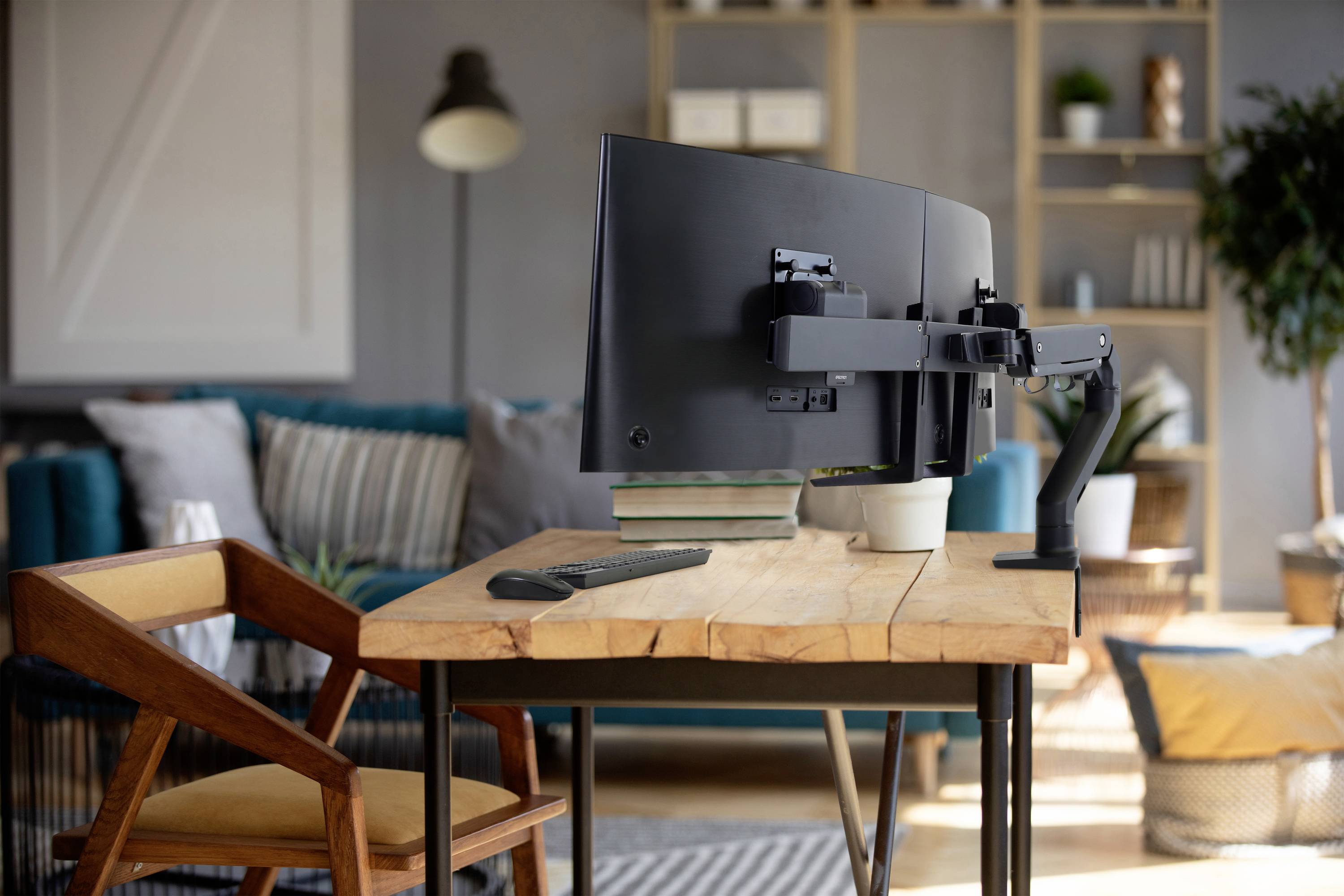 Biurko z dużym monitorem zamontowanym na ramieniu stoi w przytulnym salonie, w tle widać regały i fotel.