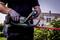 Osoba używa elektrycznych nożyc do żywopłotu, aby przycinać krzewy w ogrodzie. W tle widoczny jest dom mieszkalny.