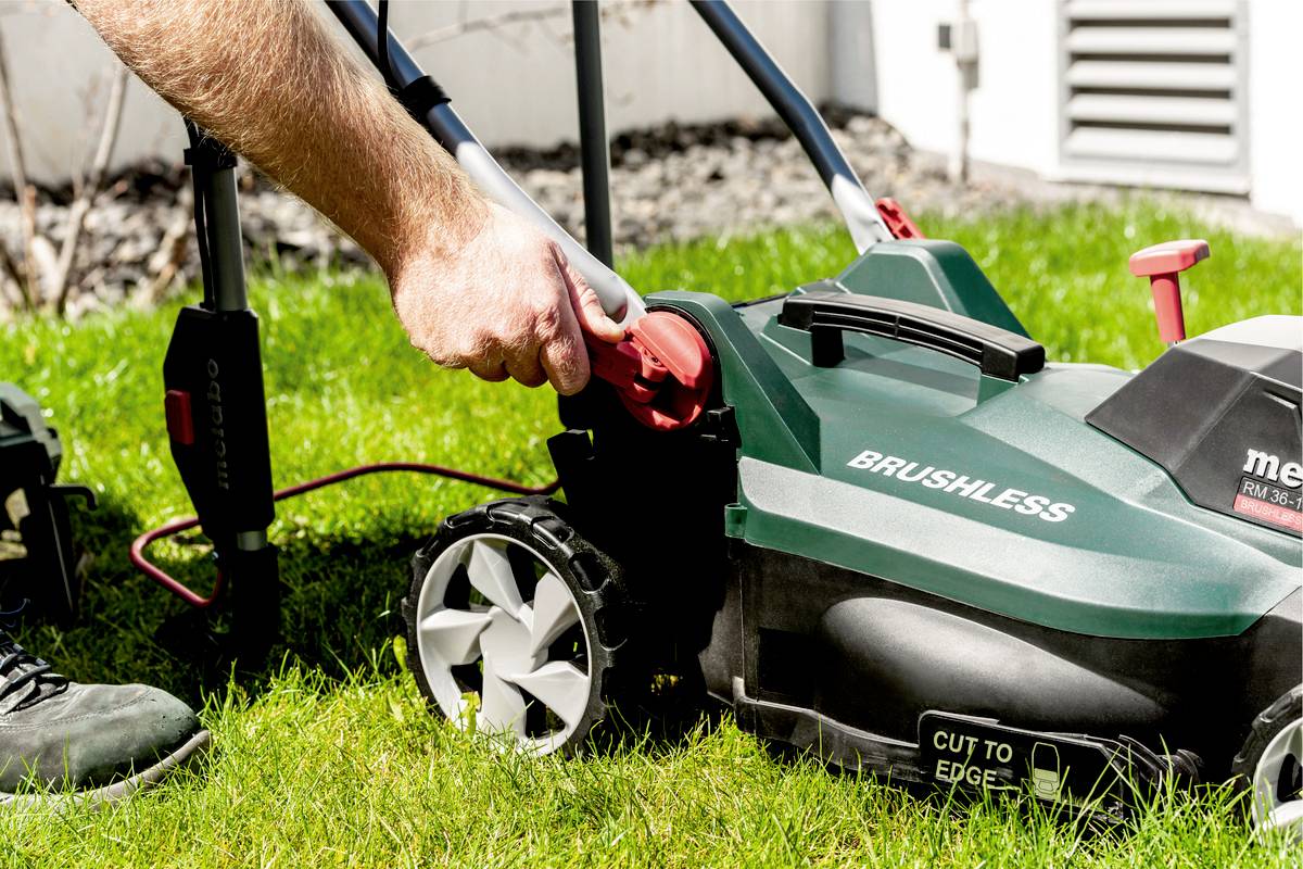 Osoba wyciąga wtyczkę elektrycznej kosiarki w ogrodzie. Kosiarka jest zielona i ma duże, srebrne koła.