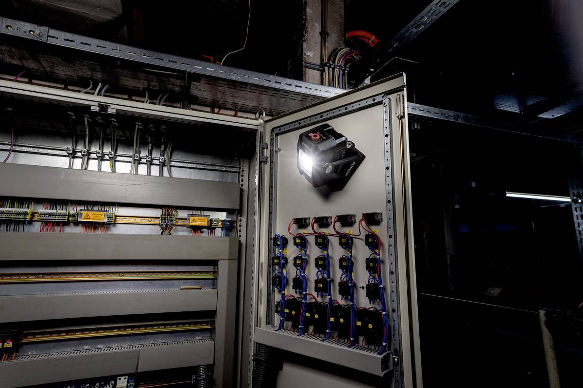 Otwarty rozdzielnik elektryczny z wieloma bezpiecznikami i wiązkami przewodów, oświetlony zamocowaną lampą, w przemysłowym otoczeniu.