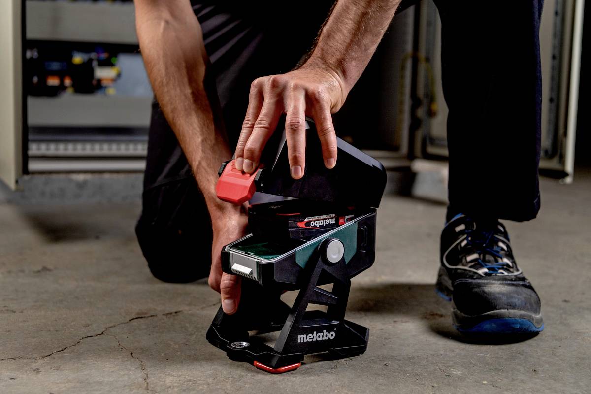 Osoba mocuje czerwone narzędzie elektryczne na stojaku na podłodze w przemysłowym otoczeniu. W tle widoczne są szafki.
