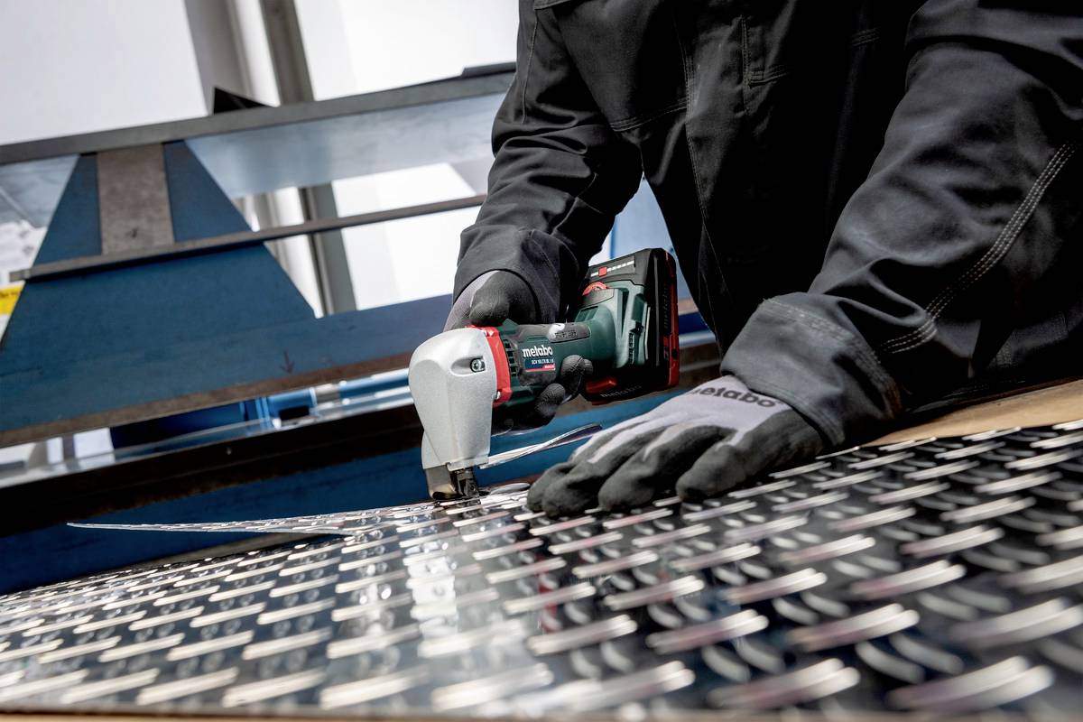 Osoba tnie blachę elektryczną maszyną tnącą w warsztacie, ubrana w rękawice i odzież ochronną.