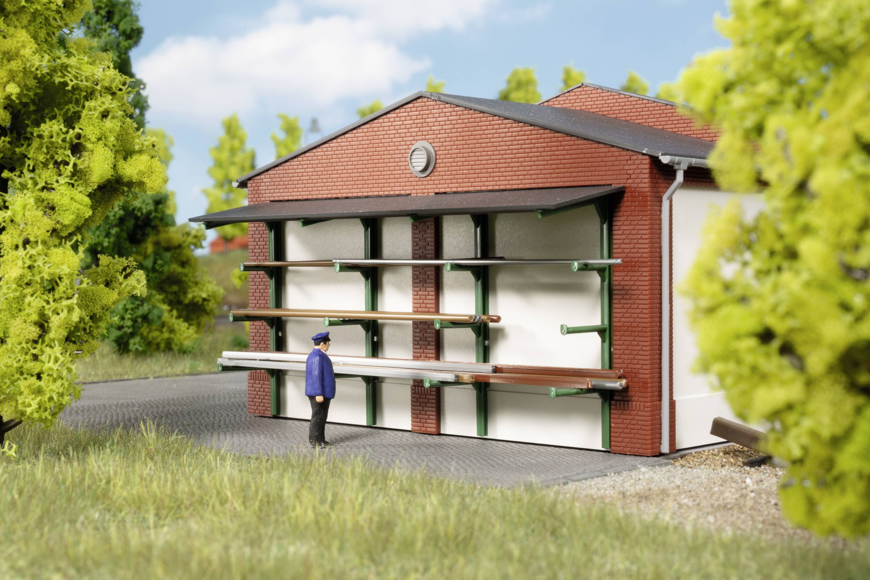 Mężczyzna w niebieskim ubraniu stoi przed dużym budynkiem z wystającymi, poziomymi elementami metalowymi. Drzewa otaczają scenę.