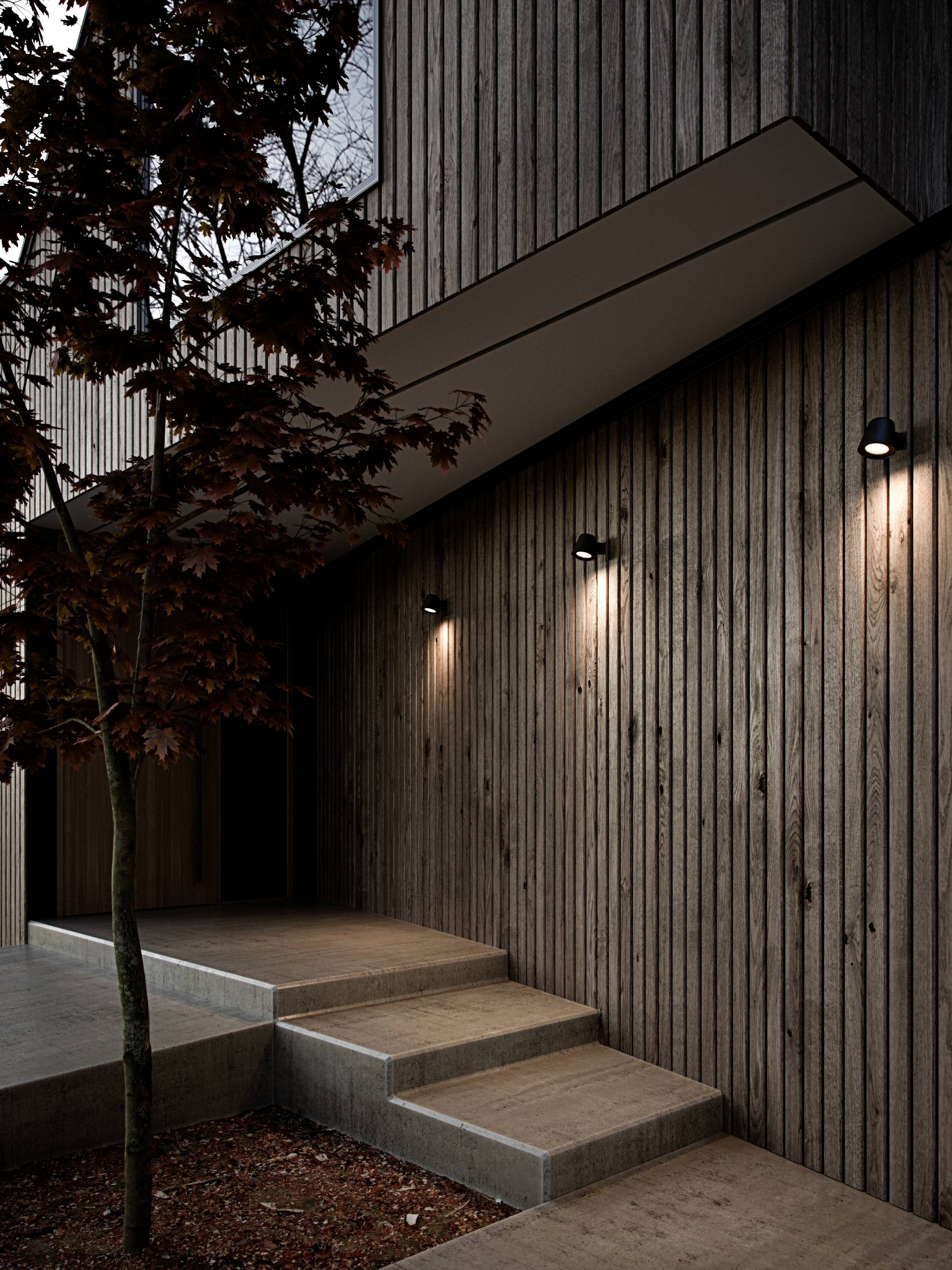 Nowoczesna drewniana fasada budynku z oświetleniem. Wąskie schody prowadzą do wejścia. Obok stoi drzewo. Nastój zmierzchu.